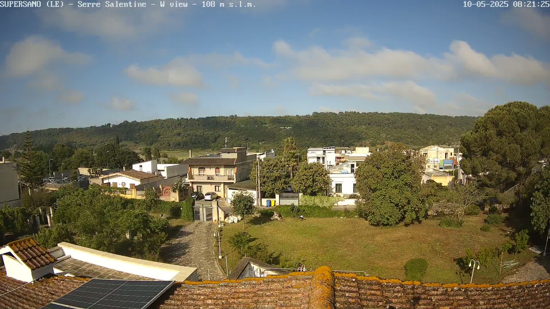 A partly sunny day reveals a view of a residential area nestled against a verdant hillside, observed from a rooftop with solar panels.