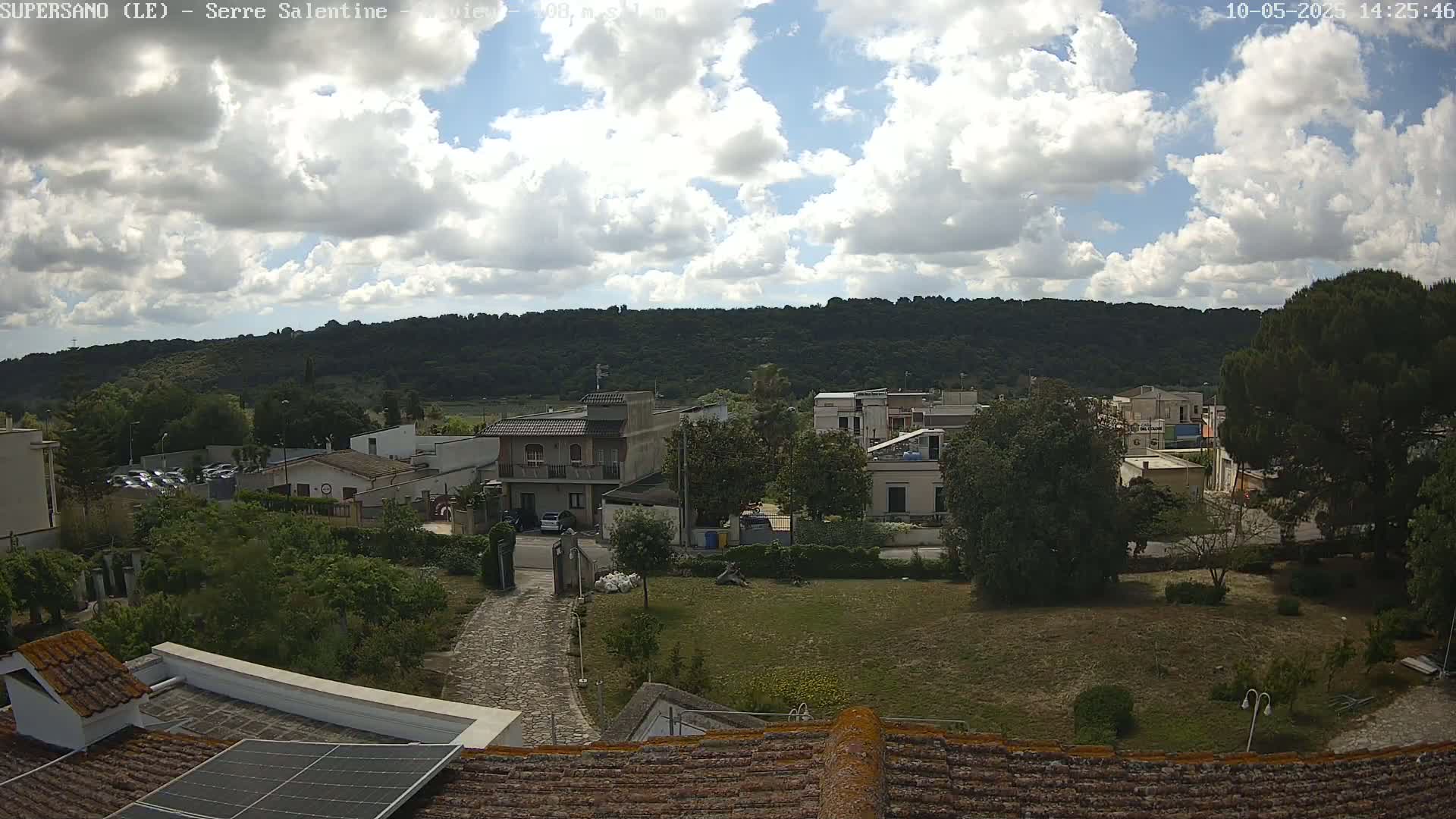 A partly cloudy day reveals a view of a residential area nestled against a verdant hillside, with a stone driveway leading to a cluster of buildings.