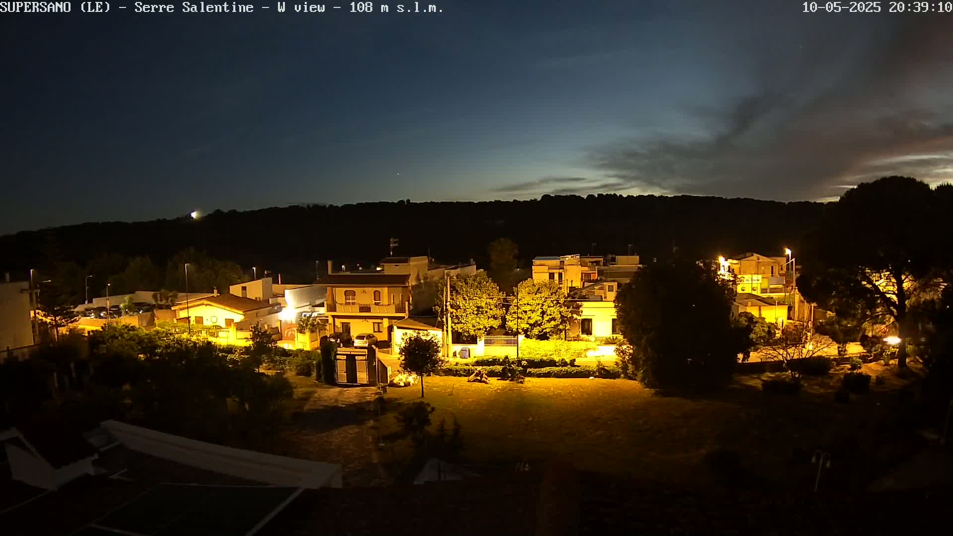 A nighttime view of a residential area illuminated by streetlights, under a mostly clear sky with a few clouds.