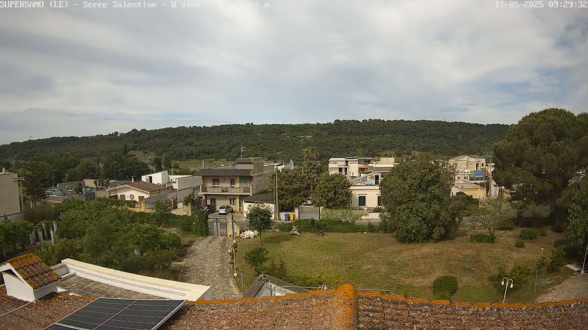 A slightly overcast day reveals a residential area nestled at the foot of a verdant hill, viewed from a rooftop with a solar panel.