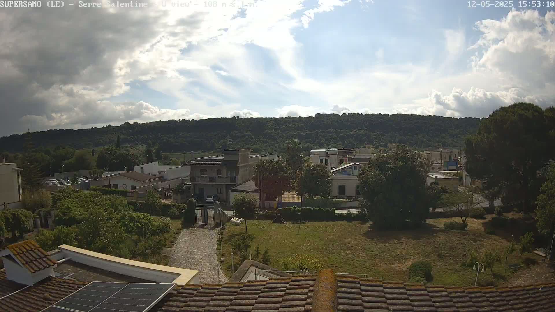 A partly cloudy sky overlooks a residential area with green lawns, trees, and buildings nestled at the base of a wooded hill.