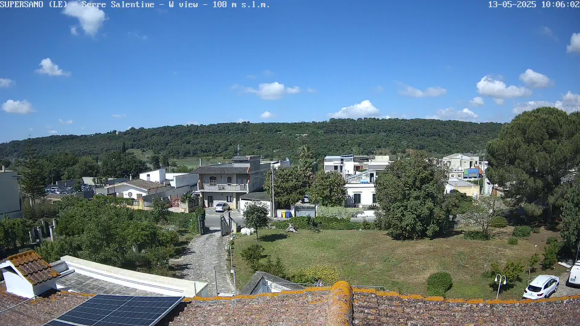 A partly sunny day reveals a residential area nestled below a tree-covered hillside, viewed from a rooftop with a solar panel.