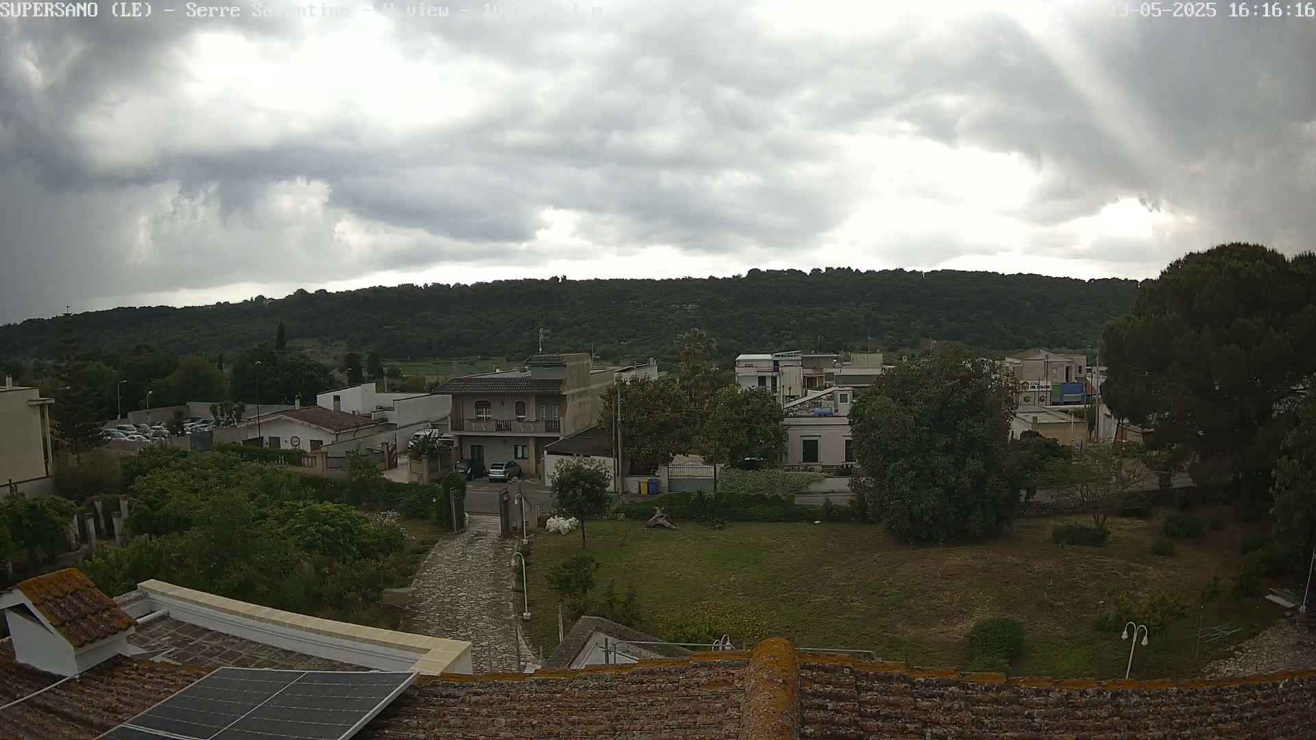 A view of a residential area with overcast skies shows a collection of houses, trees, and a stone pathway, all set against a backdrop of a tree-covered hill.