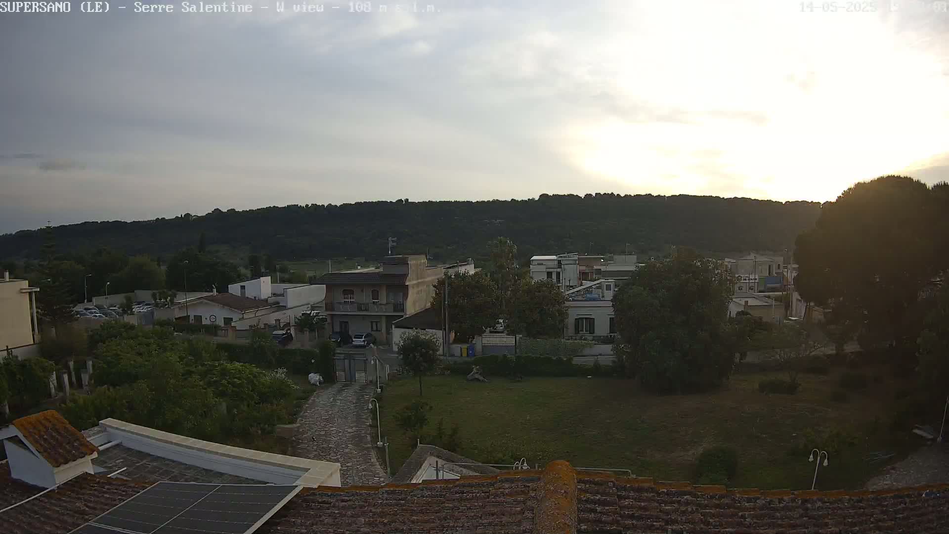 A slightly overcast sunset illuminates a residential area nestled below a tree-covered hill.