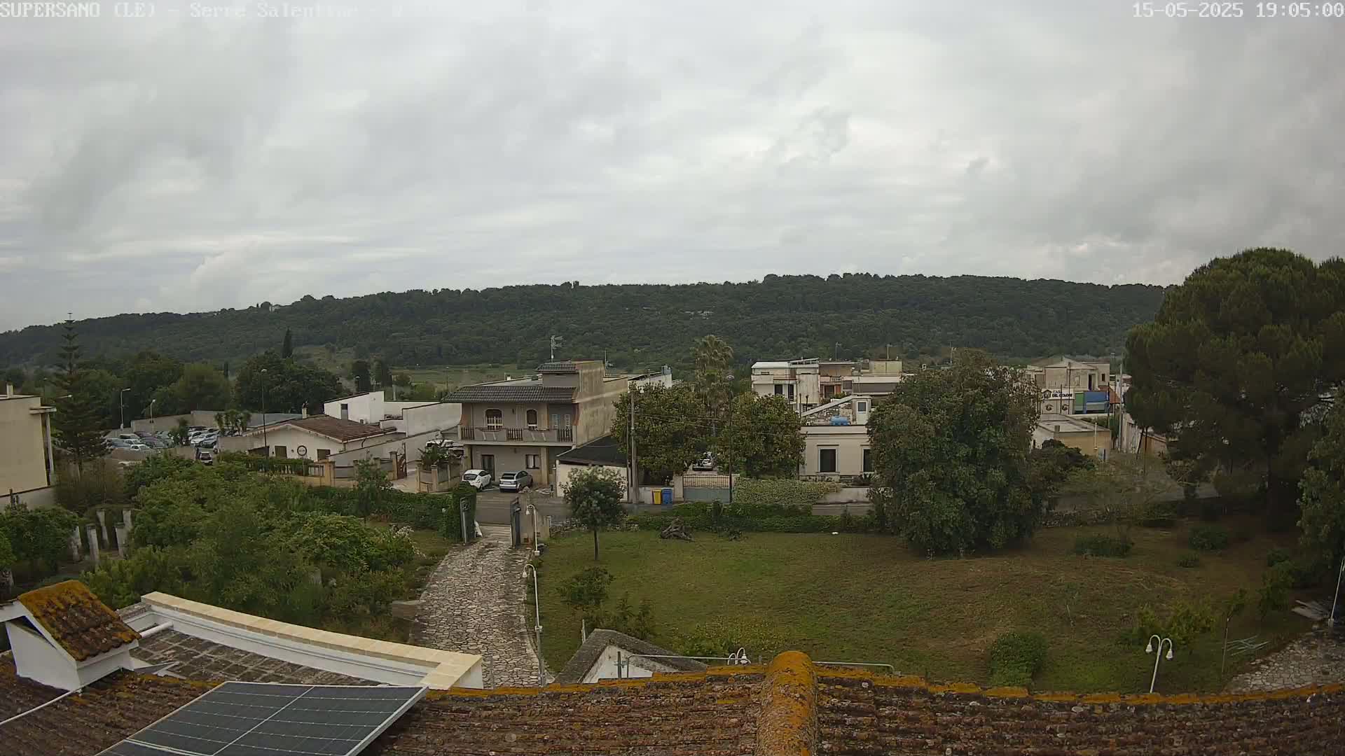 A view from a rooftop shows a residential area with overcast skies, green vegetation, and a hill in the background.