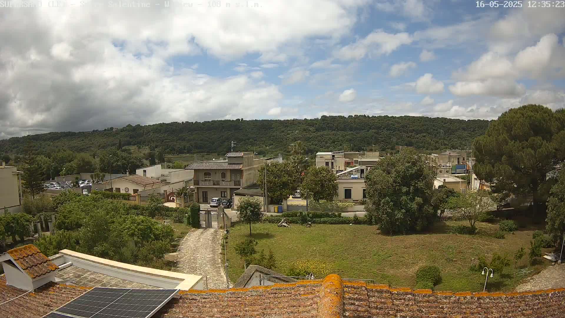 A partly cloudy day overlooks a residential area with low buildings and green vegetation, beyond which lies a tree-covered hillside.