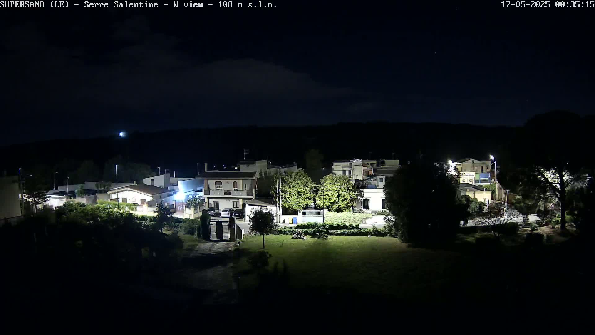 A nighttime, high-angle view shows a residential area with houses and streetlights under a mostly clear, dark sky.