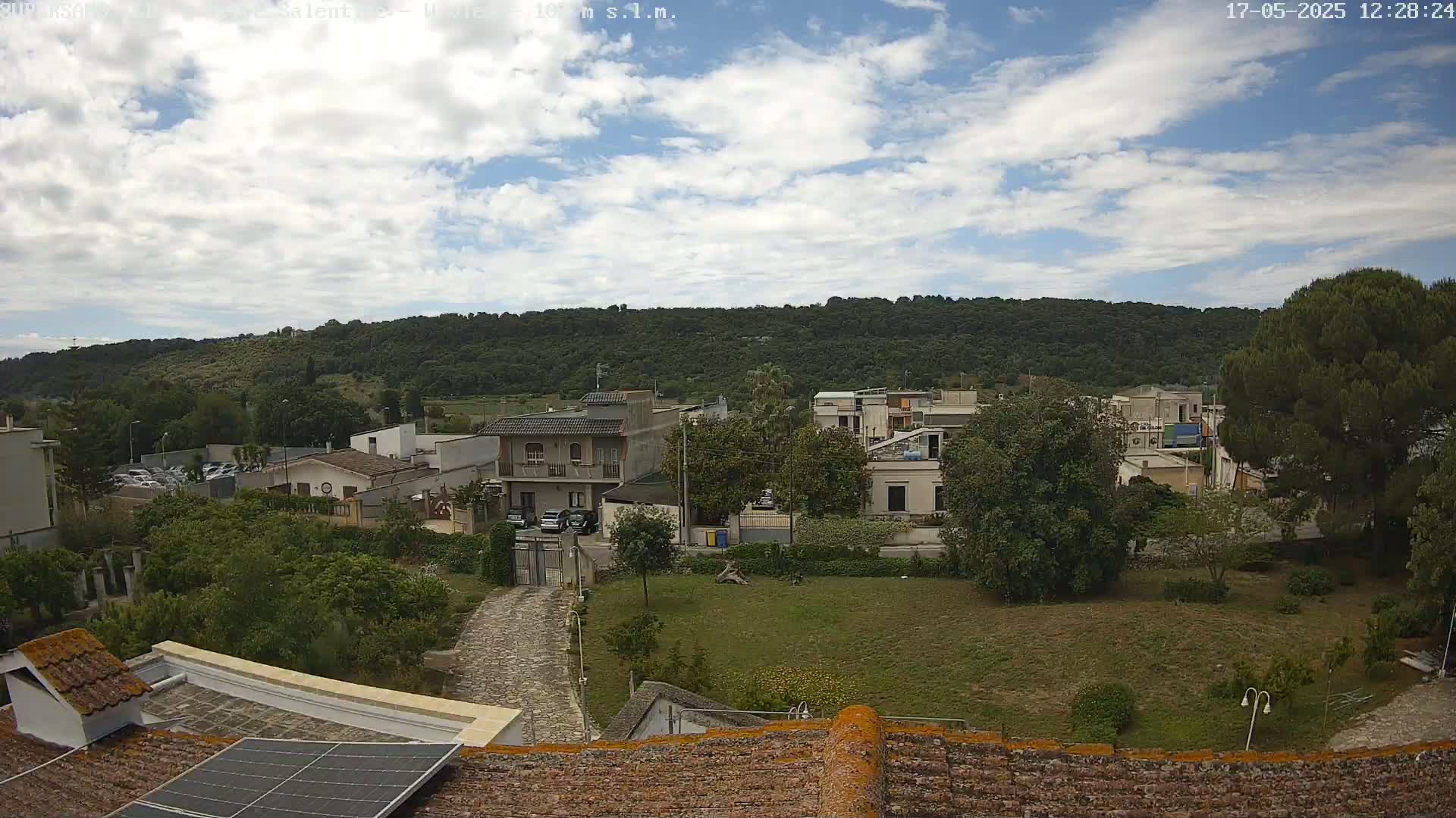 A partly cloudy sky overlooks a residential area with low buildings and green vegetation, including a hill in the background.