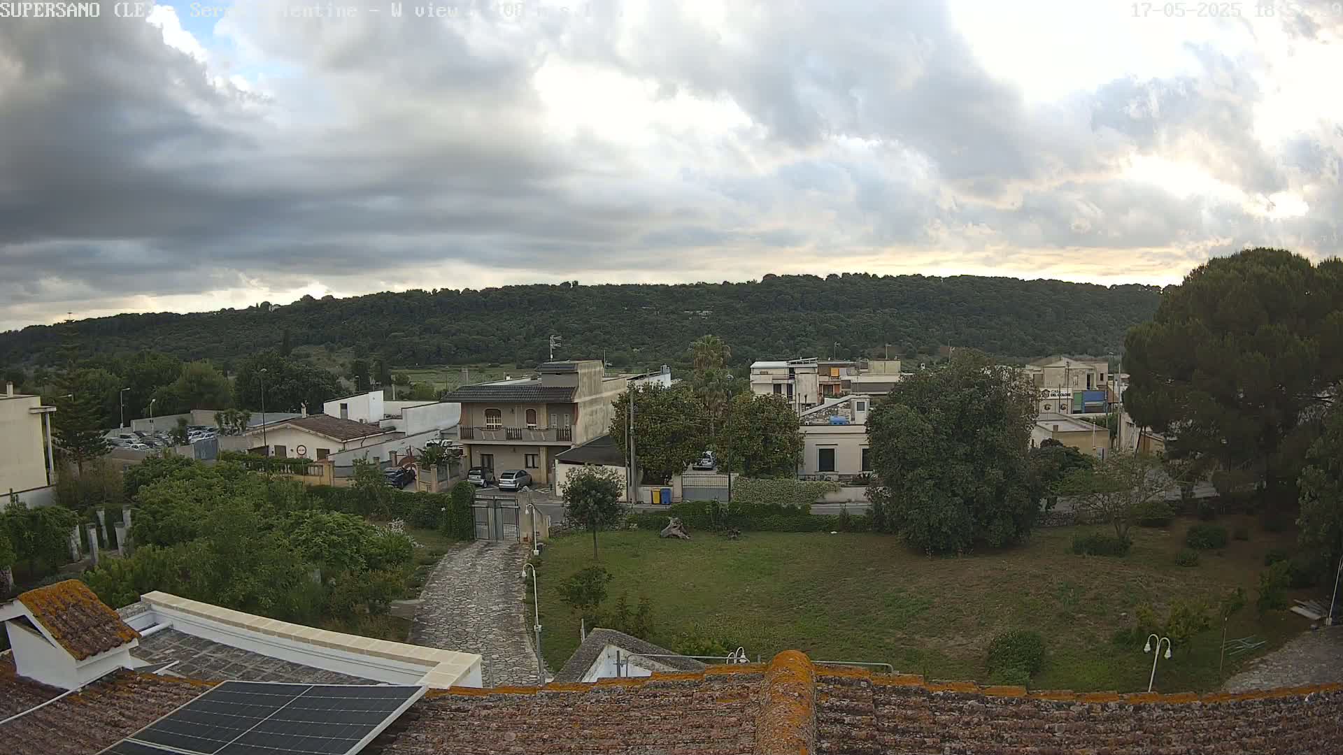 A partly cloudy sky overlooks a residential area with low-lying buildings, green lawns, and trees, situated below a tree-covered hill.