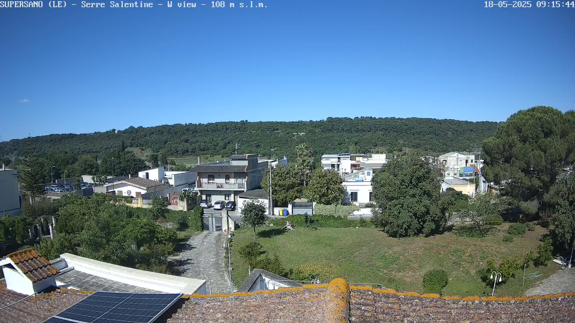 A sunny day reveals a view of a residential area nestled against a verdant hillside, seen from a rooftop with a solar panel.