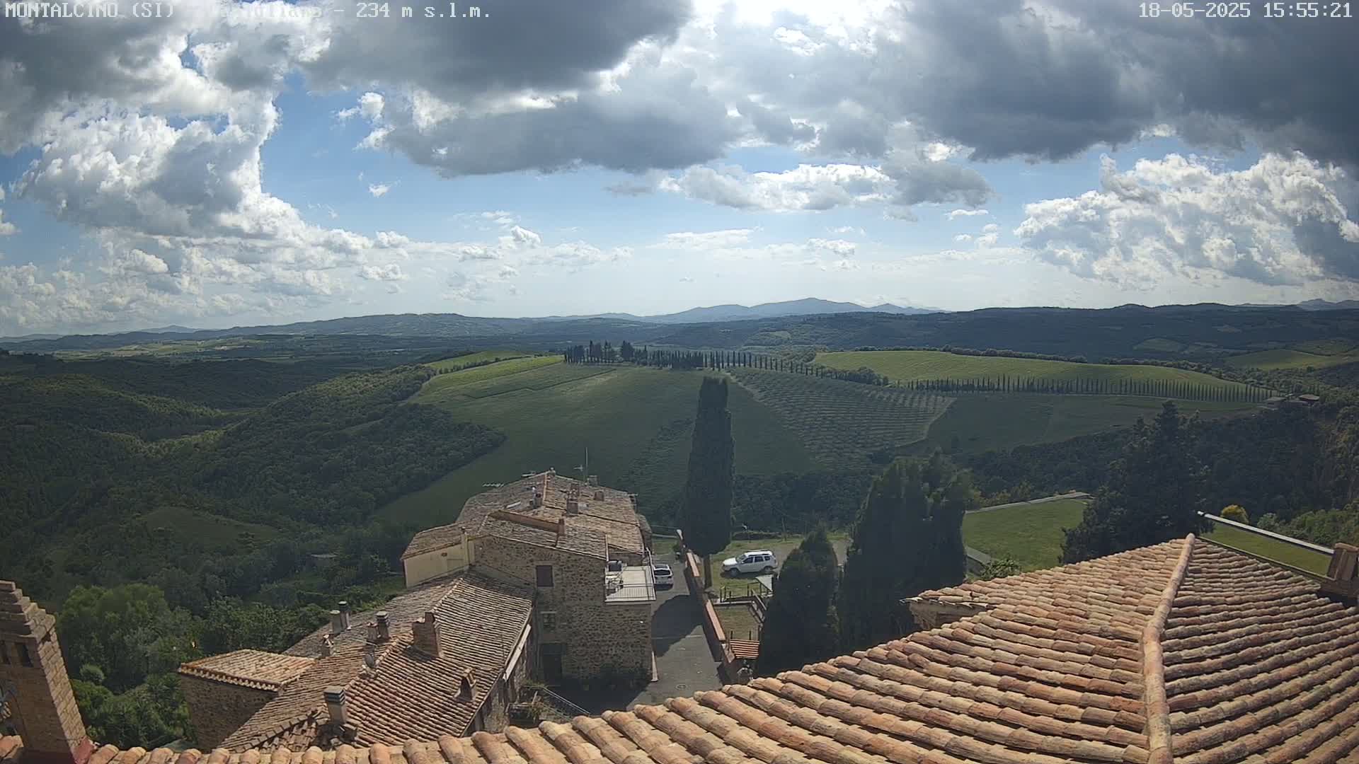 A partly cloudy sky overlooks rolling green hills dotted with cypress trees and a village with terracotta rooftops.