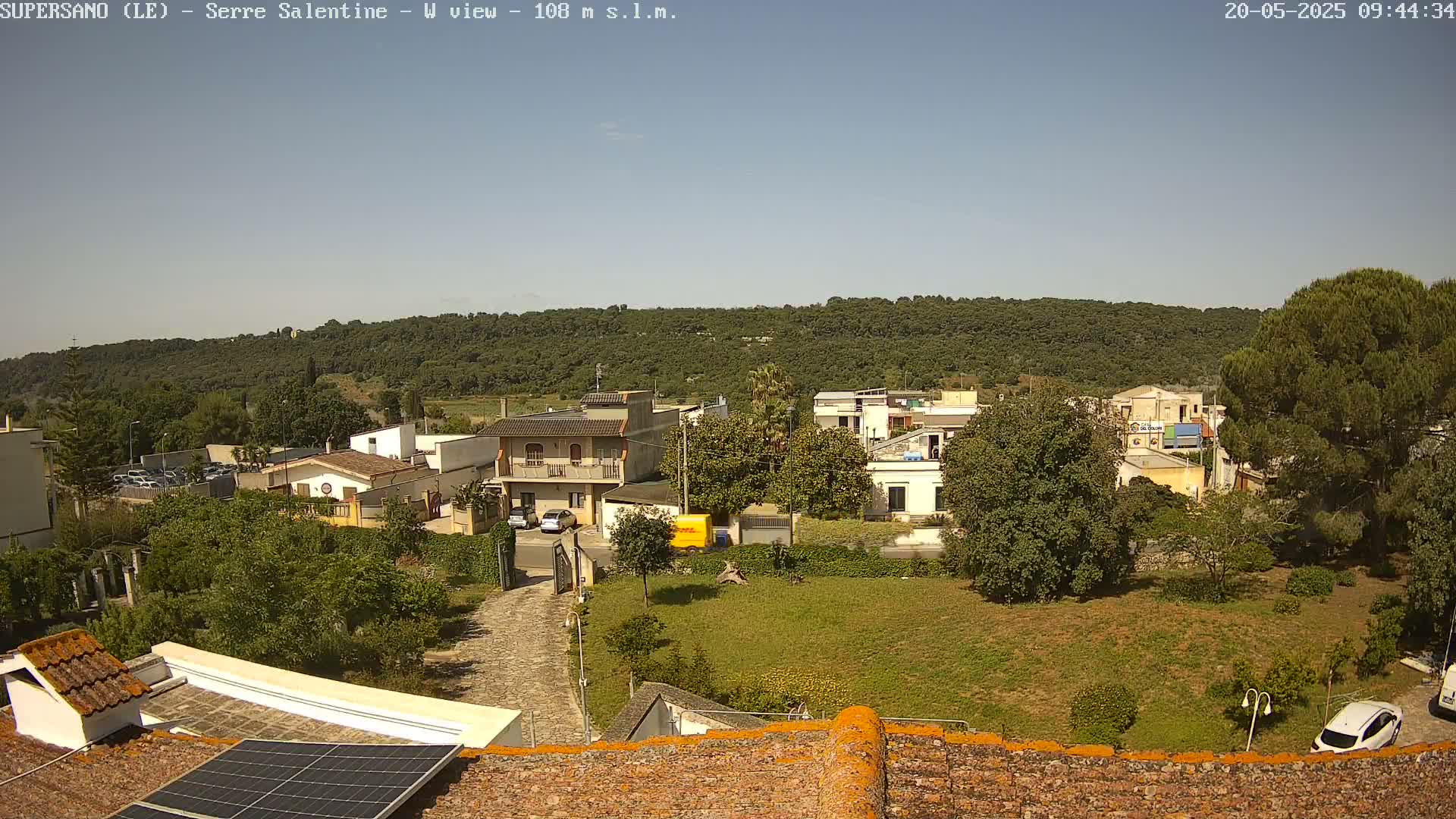 A rooftop view of a residential area on a sunny day, showing houses, trees, and a grassy yard, with a hill covered in trees in the background.