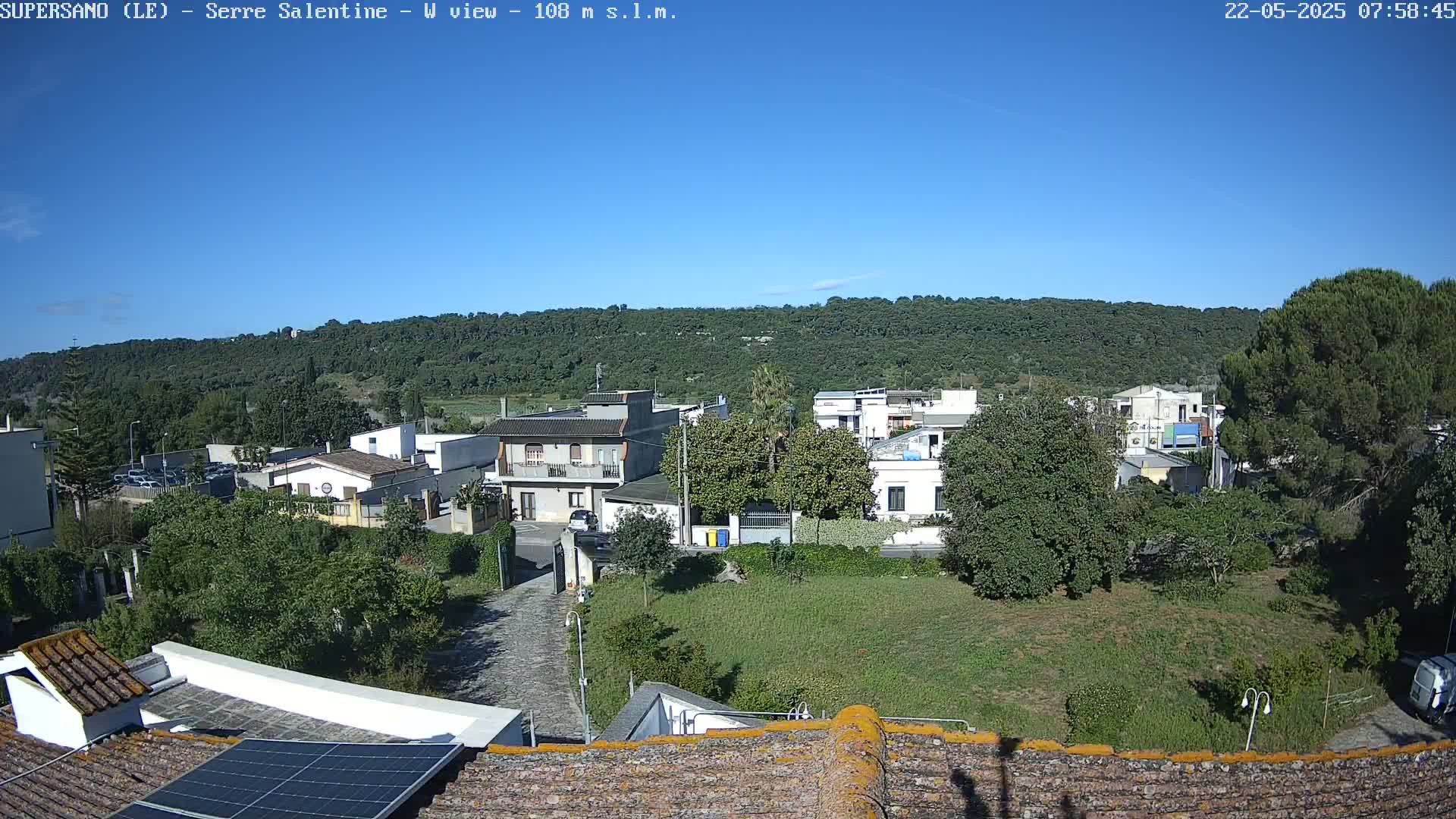 A clear, sunny day reveals a residential area nestled below a tree-covered hill, viewed from a rooftop with solar panels.