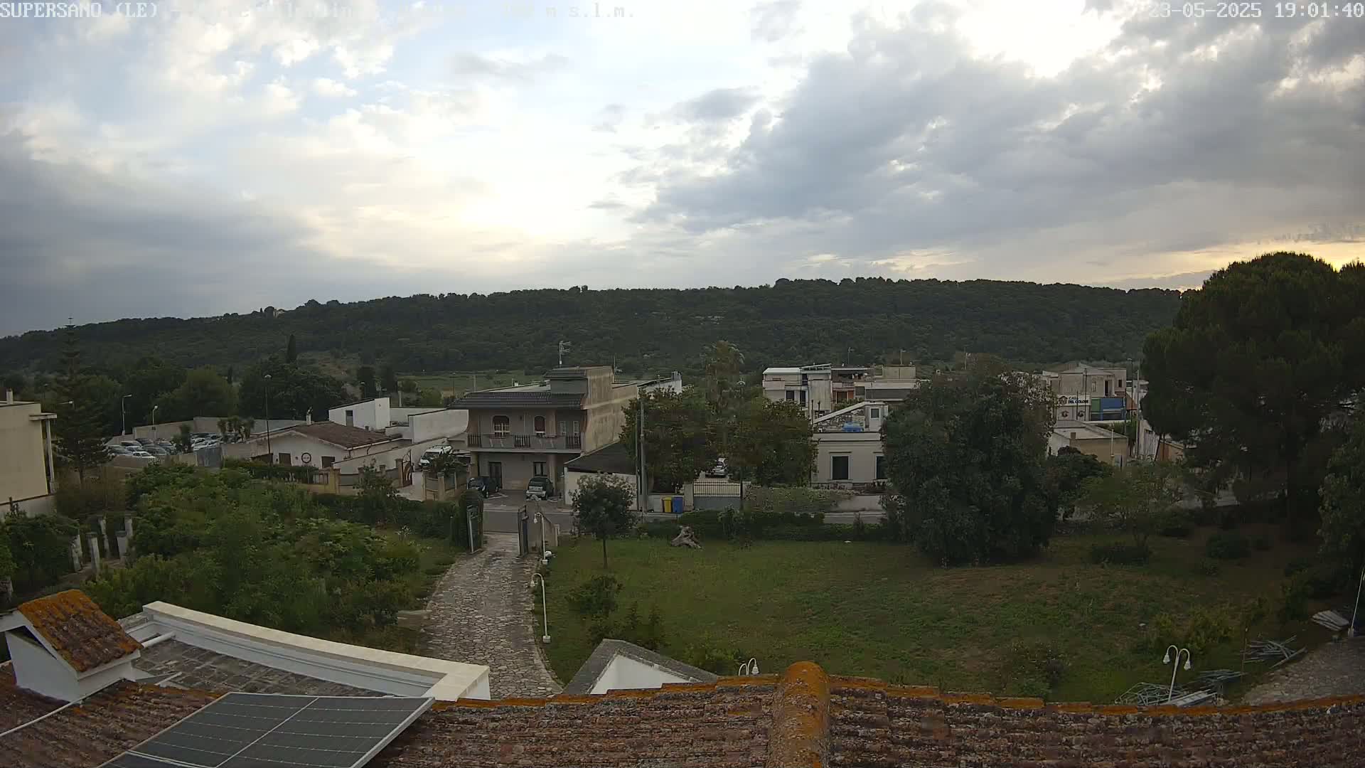 A cloudy evening view of a residential area nestled against a verdant hillside, seen from a rooftop with a solar panel.