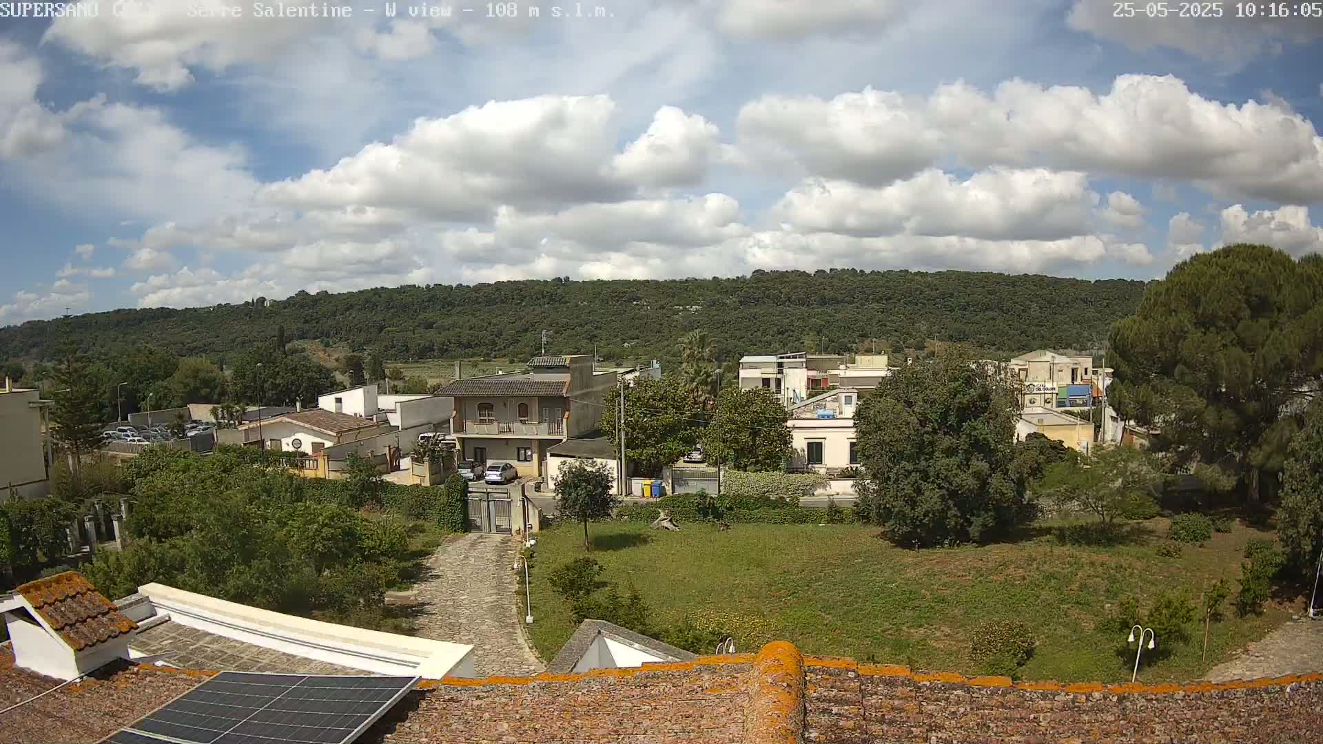A partly cloudy day reveals a view of a residential area nestled against a verdant hillside, as seen from a rooftop with a solar panel.