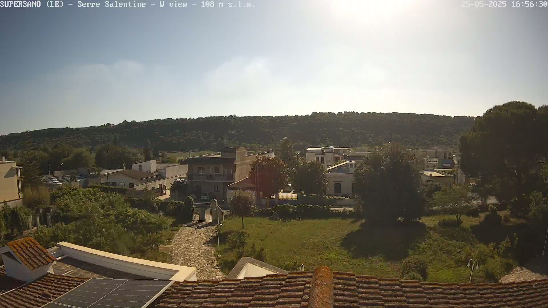 A sunny day reveals a residential area nestled at the base of a verdant hill, seen from a rooftop with solar panels.