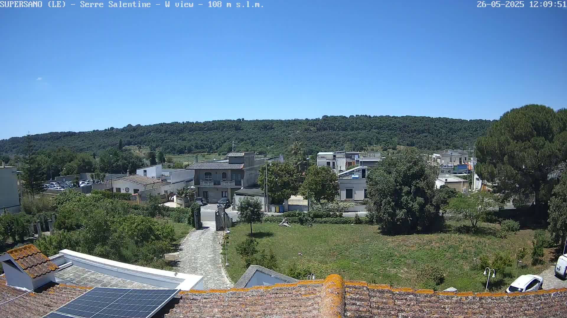 A sunny day reveals a residential area nestled at the foot of a verdant hill, with a view overlooking rooftops and  trees.