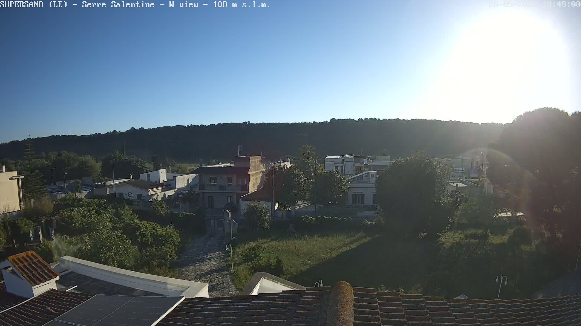 A sunny, clear day reveals a village nestled against a hillside, viewed from a rooftop with a solar panel.
