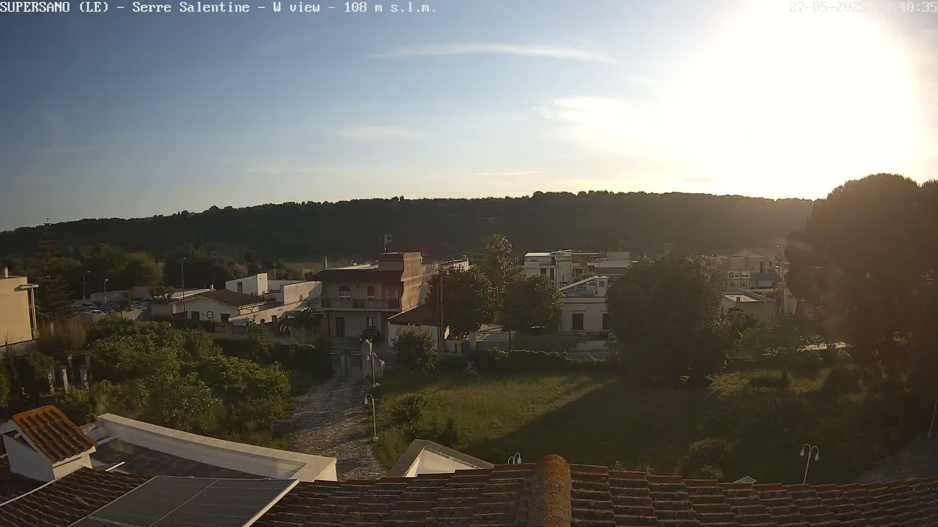 A sunlit view of a small town nestled among green hills, with rooftops and trees in the foreground.