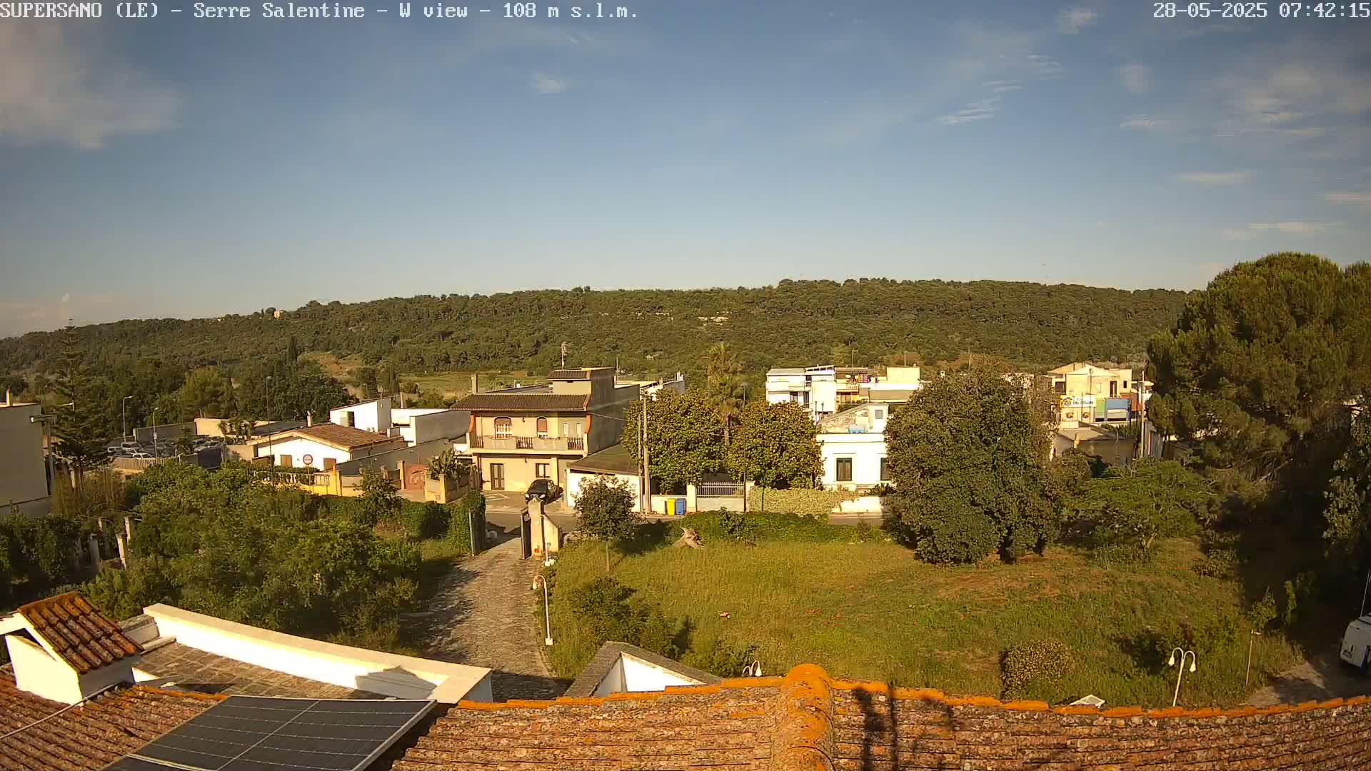 A mostly sunny day reveals a residential area with low buildings and a tree-covered hill in the background.