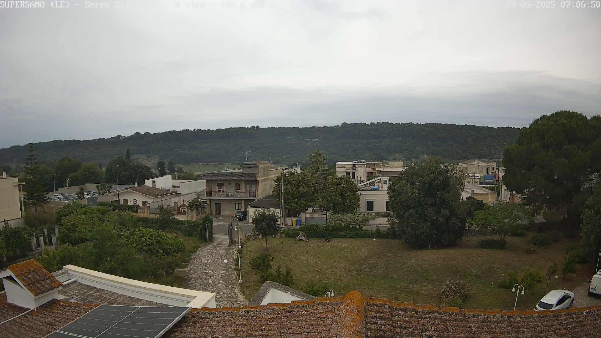 A cloudy day reveals a residential area nestled against a verdant hillside, viewed from a rooftop with a solar panel.