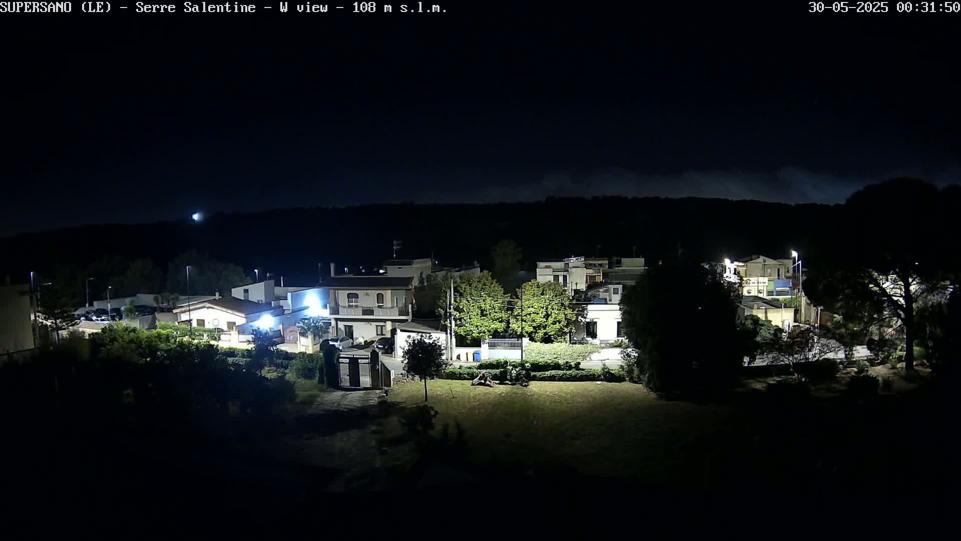 A nighttime view of a residential area with houses illuminated, under a dark sky with some cloud cover.