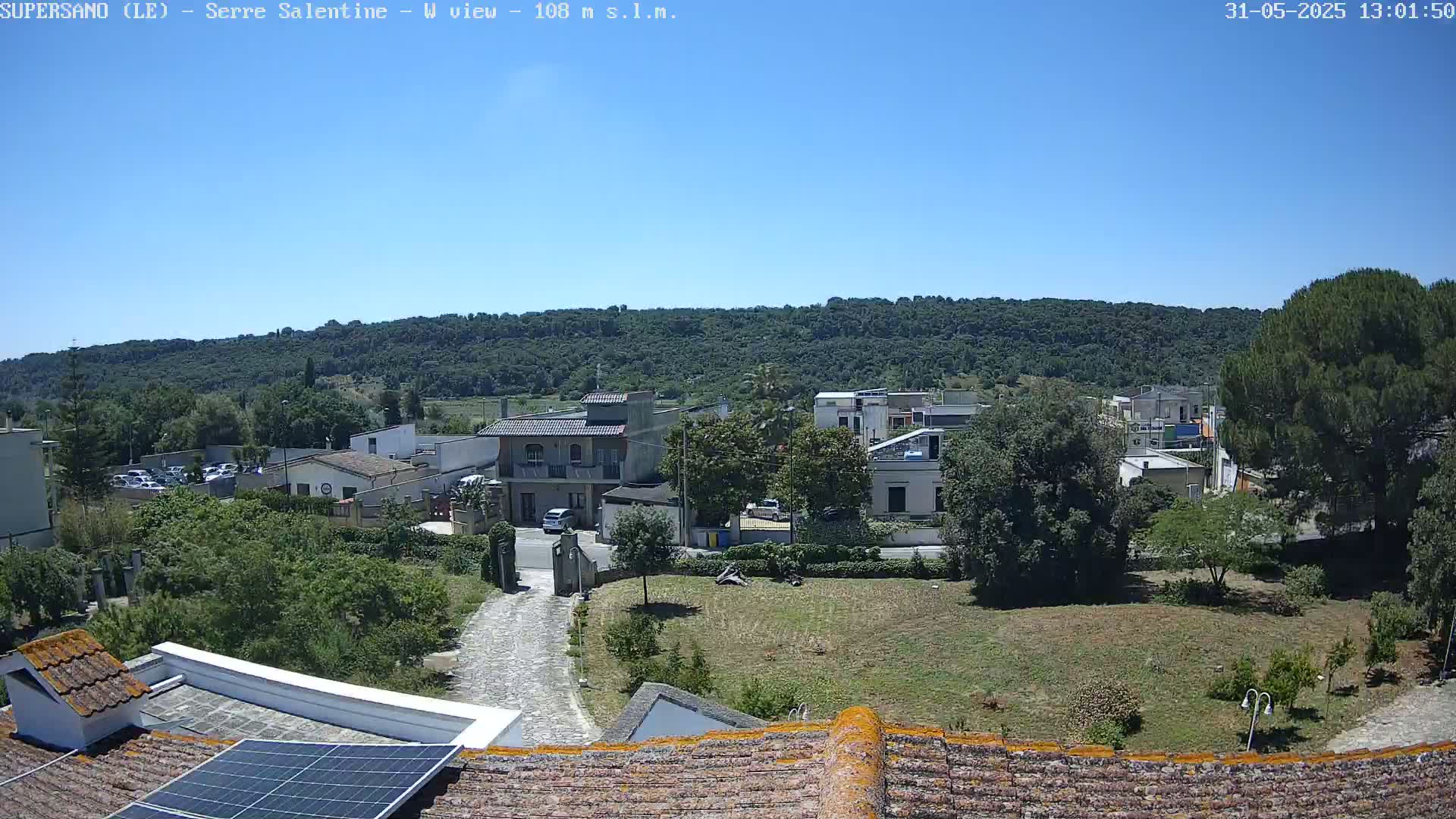 A clear, sunny day reveals a view of a small town nestled against a verdant hillside, seen from a rooftop with a solar panel.