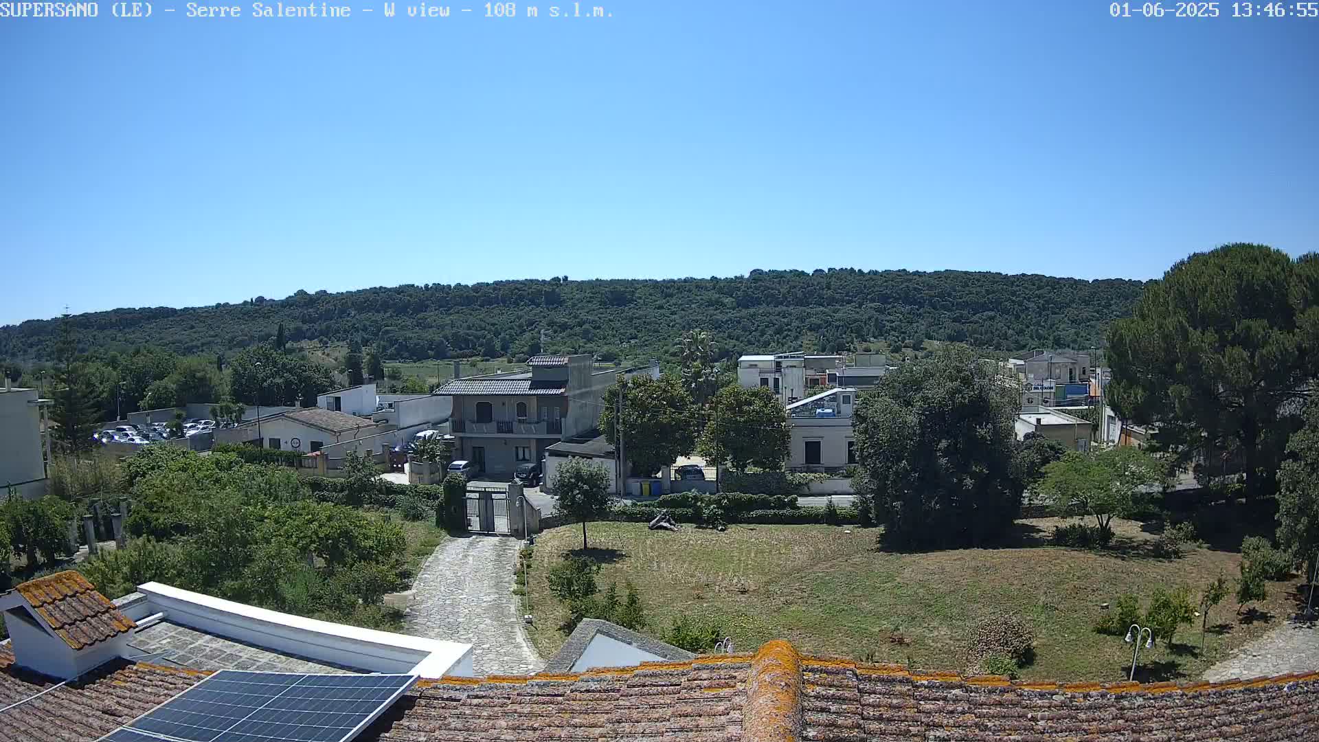 A clear, sunny day reveals a residential area nestled beside a large, verdant hill, viewed from a rooftop with a solar panel.