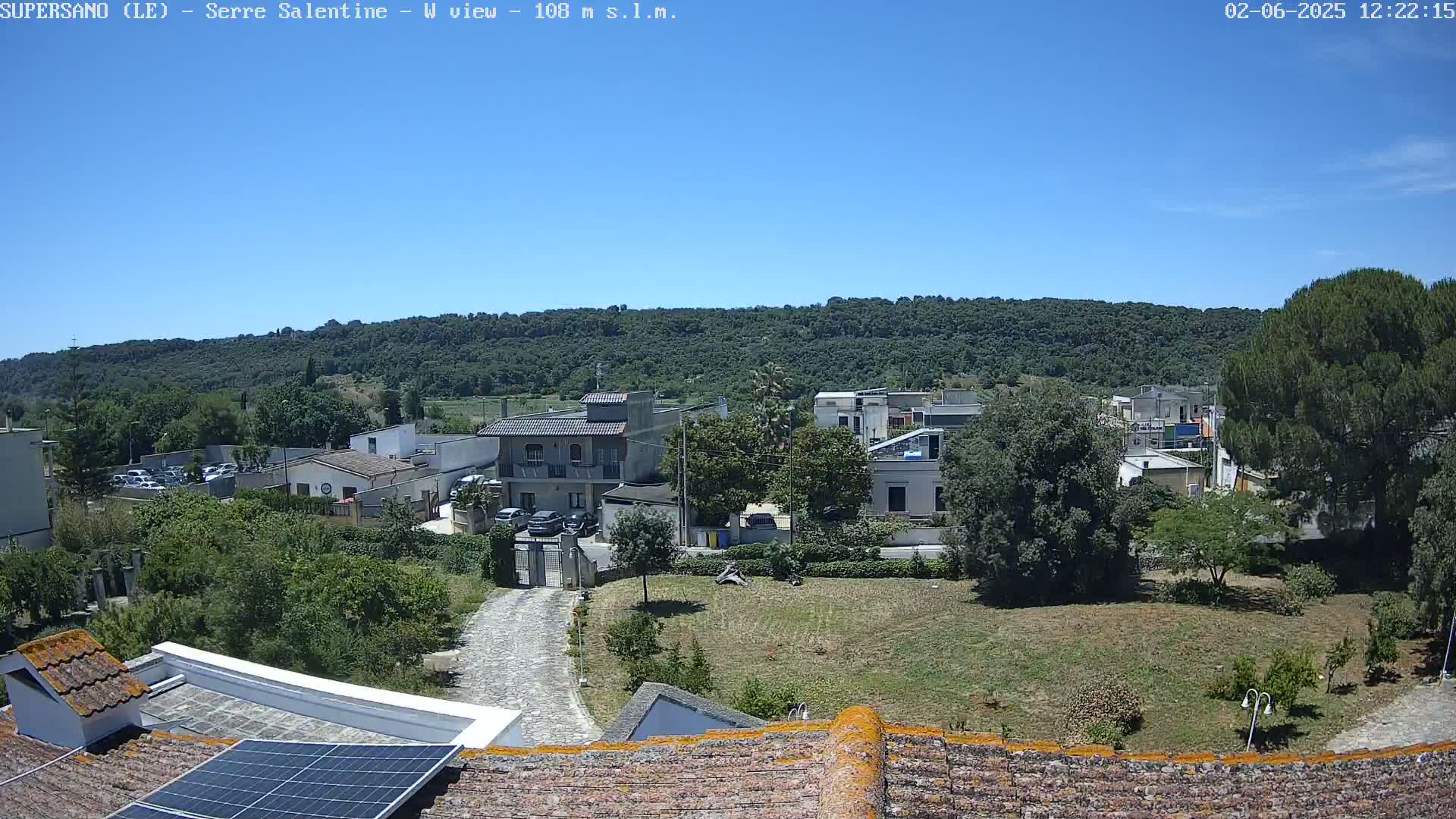A sunny day reveals a residential area nestled beside a verdant hill, viewed from a rooftop with solar panels.