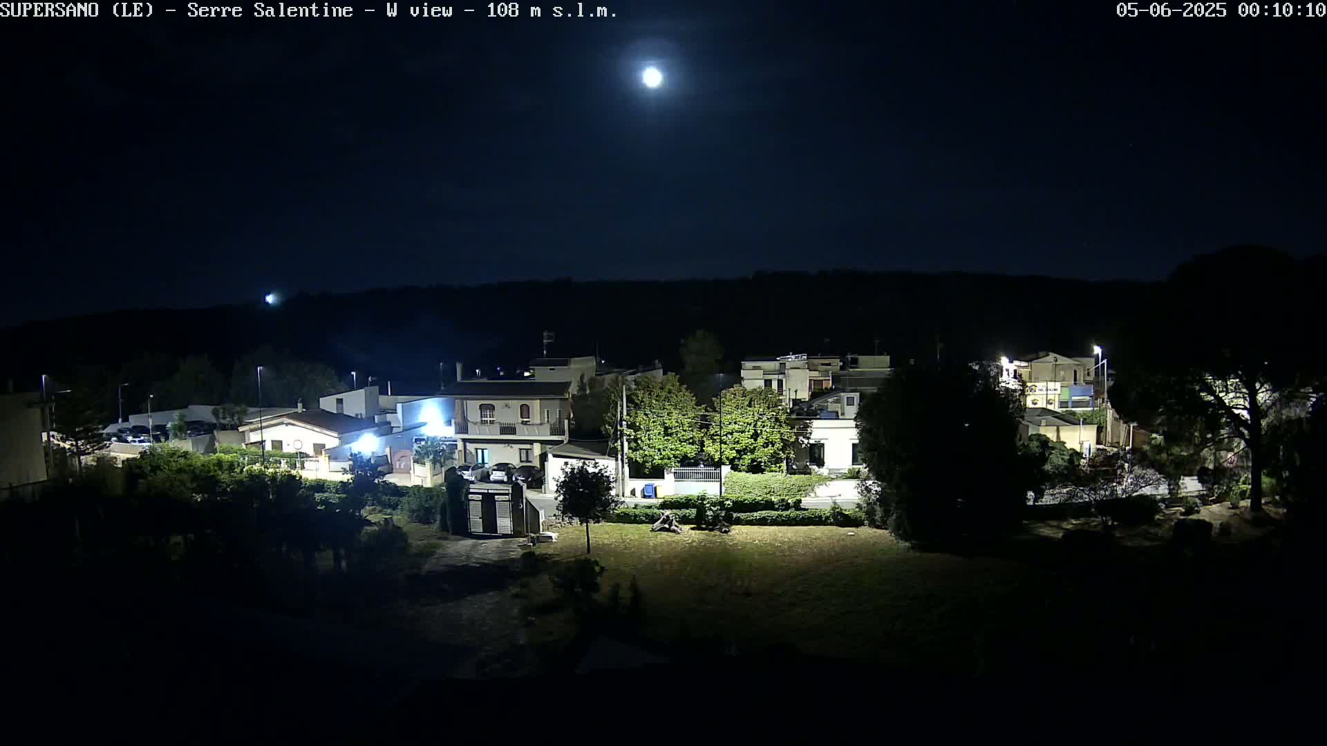 A brightly lit moon illuminates a residential area at night, revealing houses, trees, and parked cars.