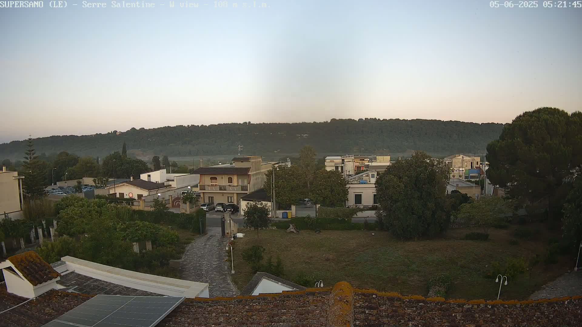A view from a rooftop shows a residential area with low buildings and trees under a clear, light-grey early morning sky.