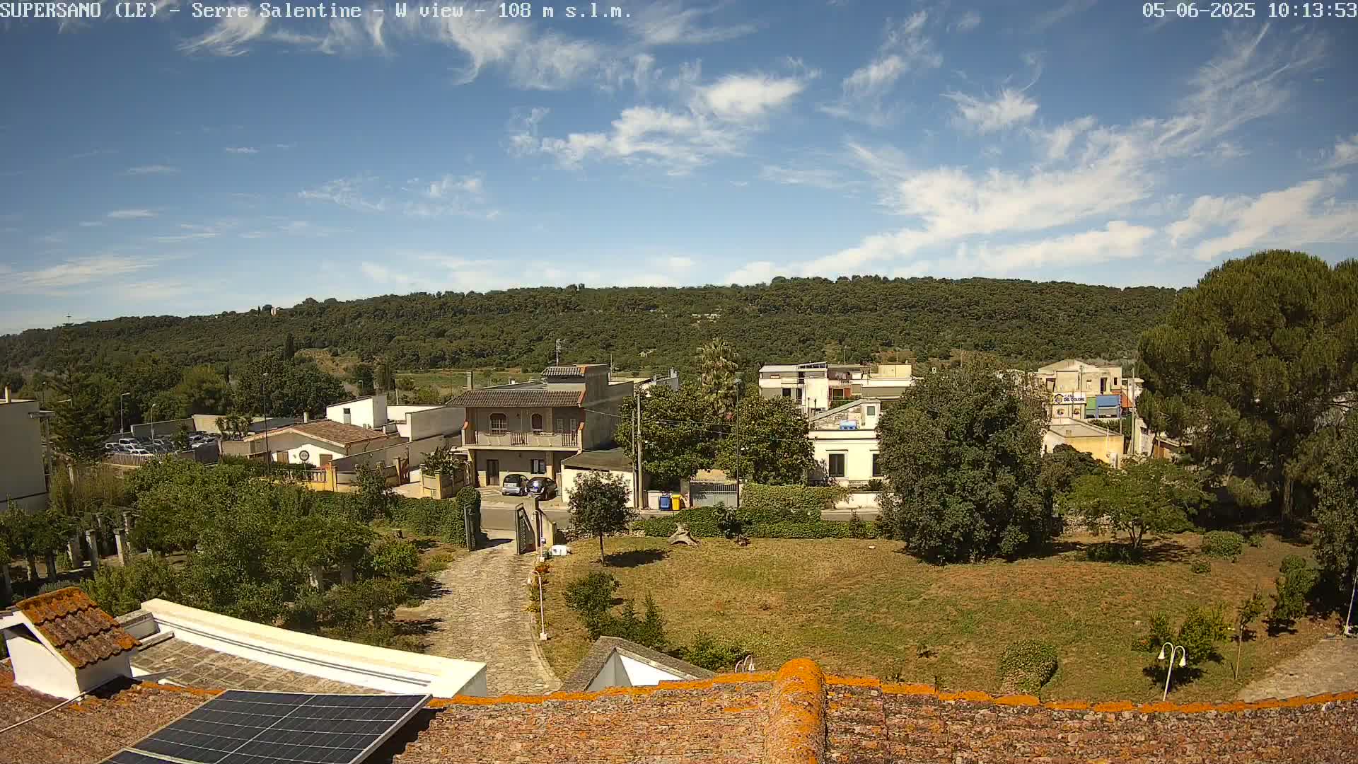 A partly sunny day reveals a residential area nestled against a verdant hillside, viewed from a rooftop with a solar panel.