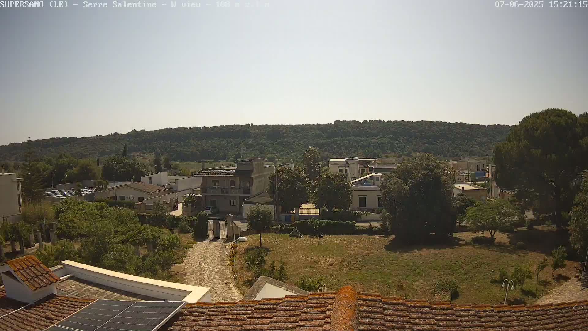 A sunny view from a rooftop shows a residential area with houses, trees, and a paved driveway leading to a gated entrance, all situated before a verdant hillside.