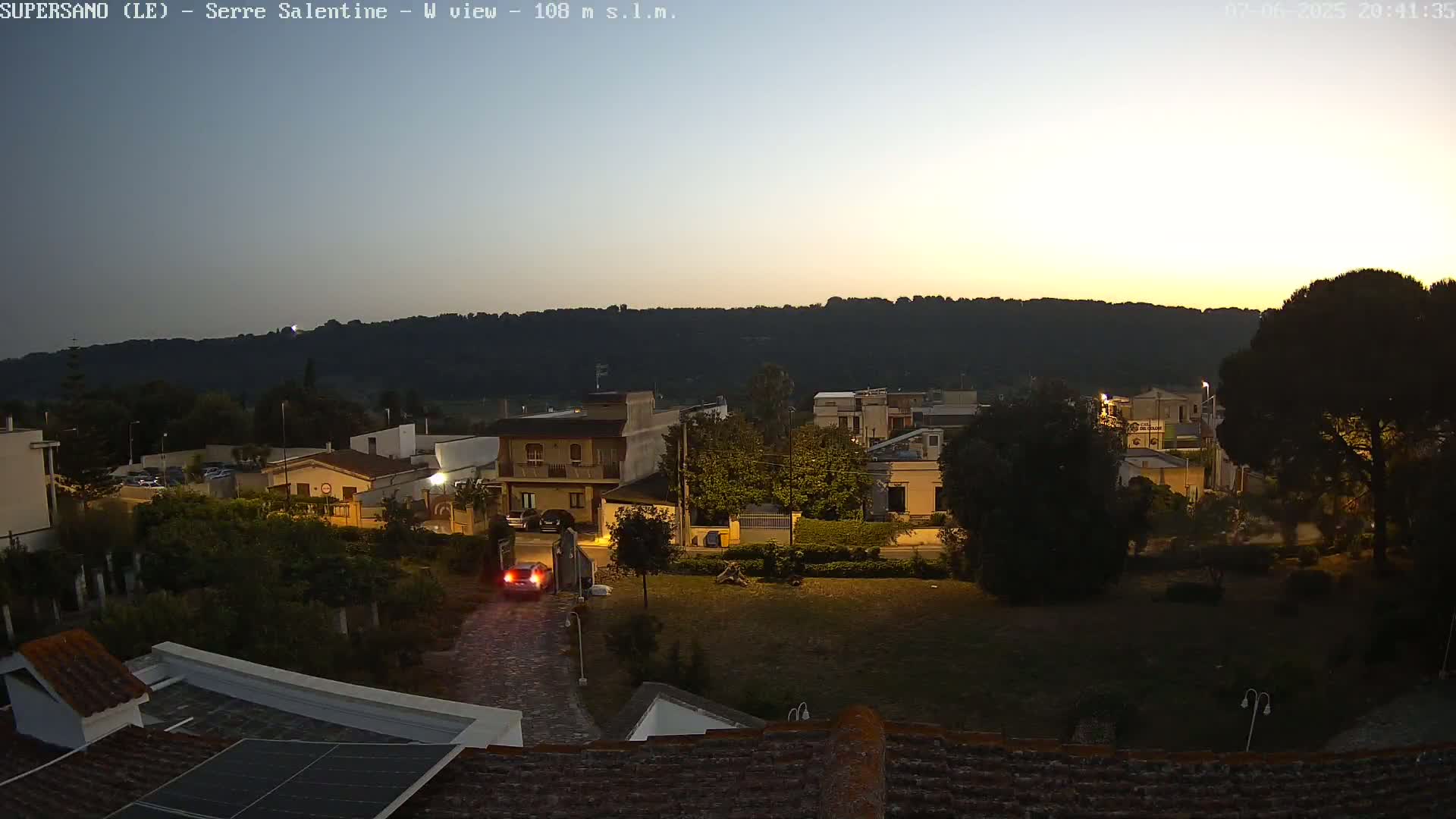 A twilight view of a small town nestled against a hill, with a few streetlights illuminating the area and a car visible on a roadway.