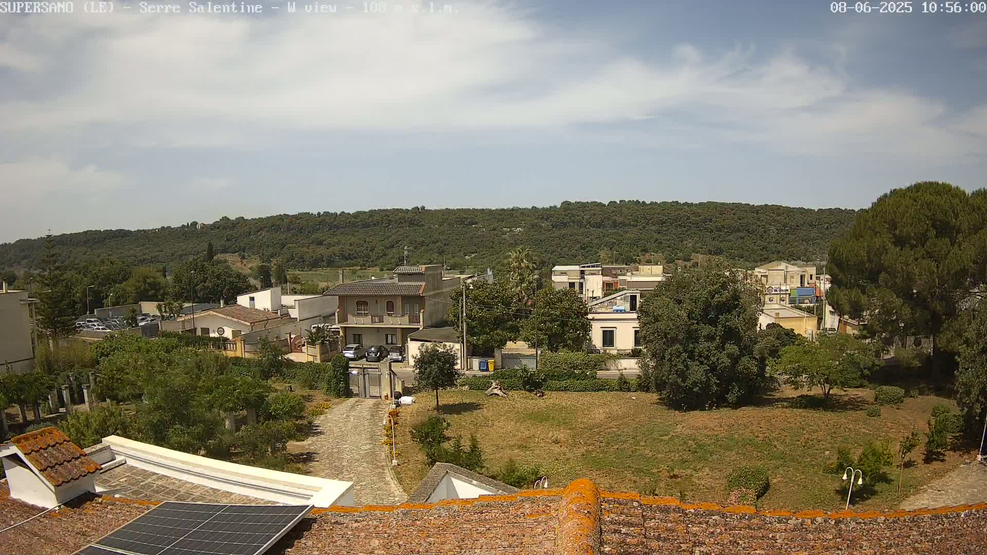 A partly sunny day reveals a residential area nestled below a verdant hill, with buildings, trees, and a paved pathway visible from a rooftop vantage point.