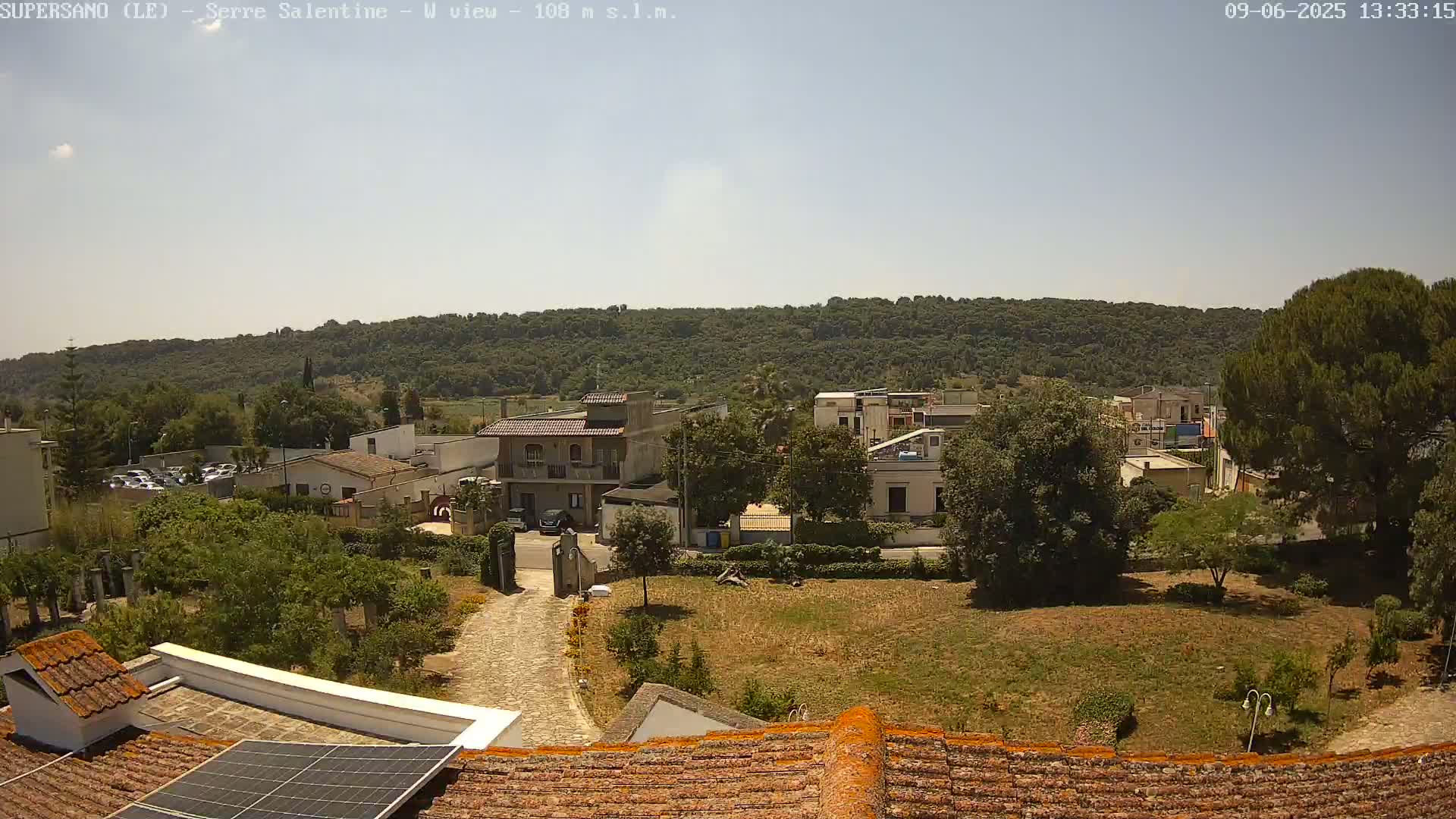 A view from a rooftop shows a village nestled amongst green hills under a clear, sunny sky.