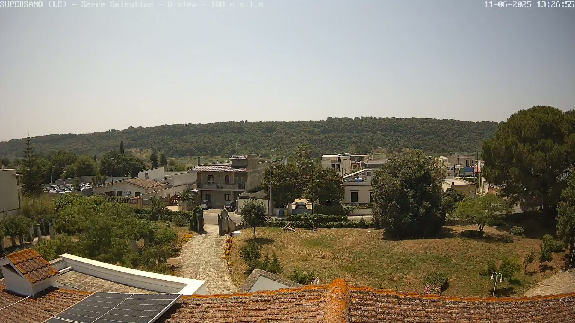 A sunny view of a small town nestled below a tree-covered hill, seen from a rooftop with solar panels.