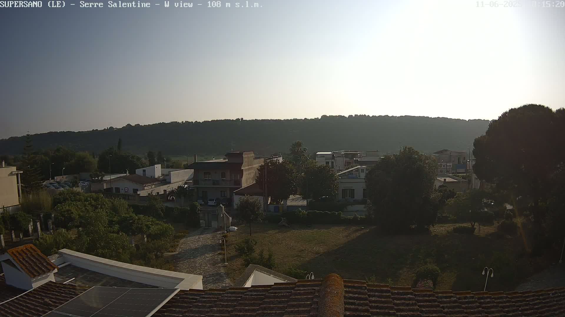 A mostly sunny view overlooking a town nestled at the base of a tree-covered hill.