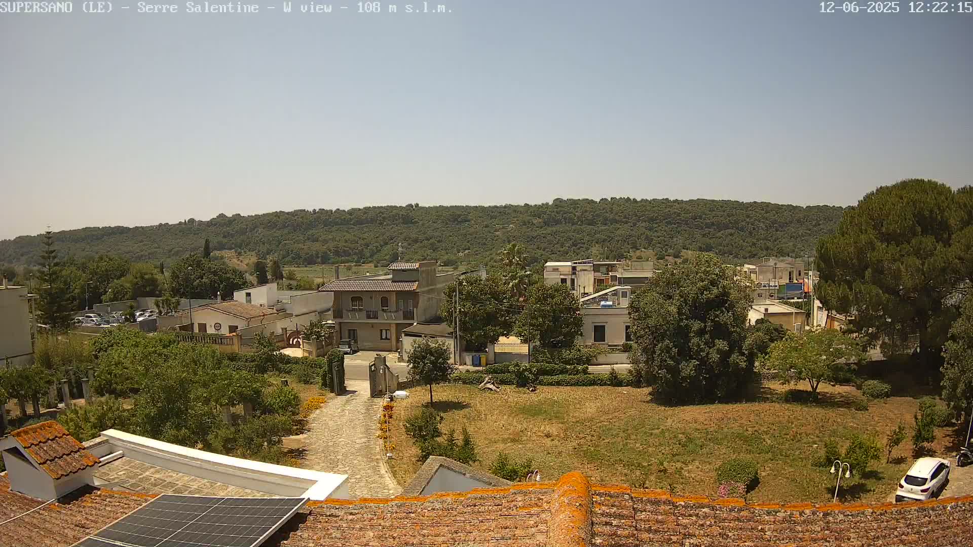 A sunlit view of a residential area with low buildings and green vegetation, hills covered in trees in the background, under a clear sky.