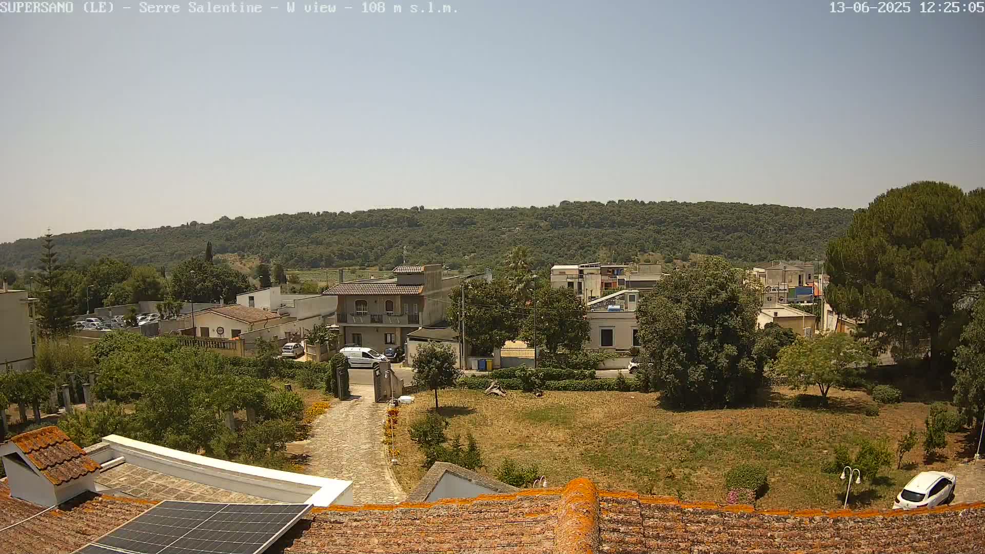 A clear, sunny day reveals a residential area nestled below a verdant hillside, with a paved driveway and a few cars visible.