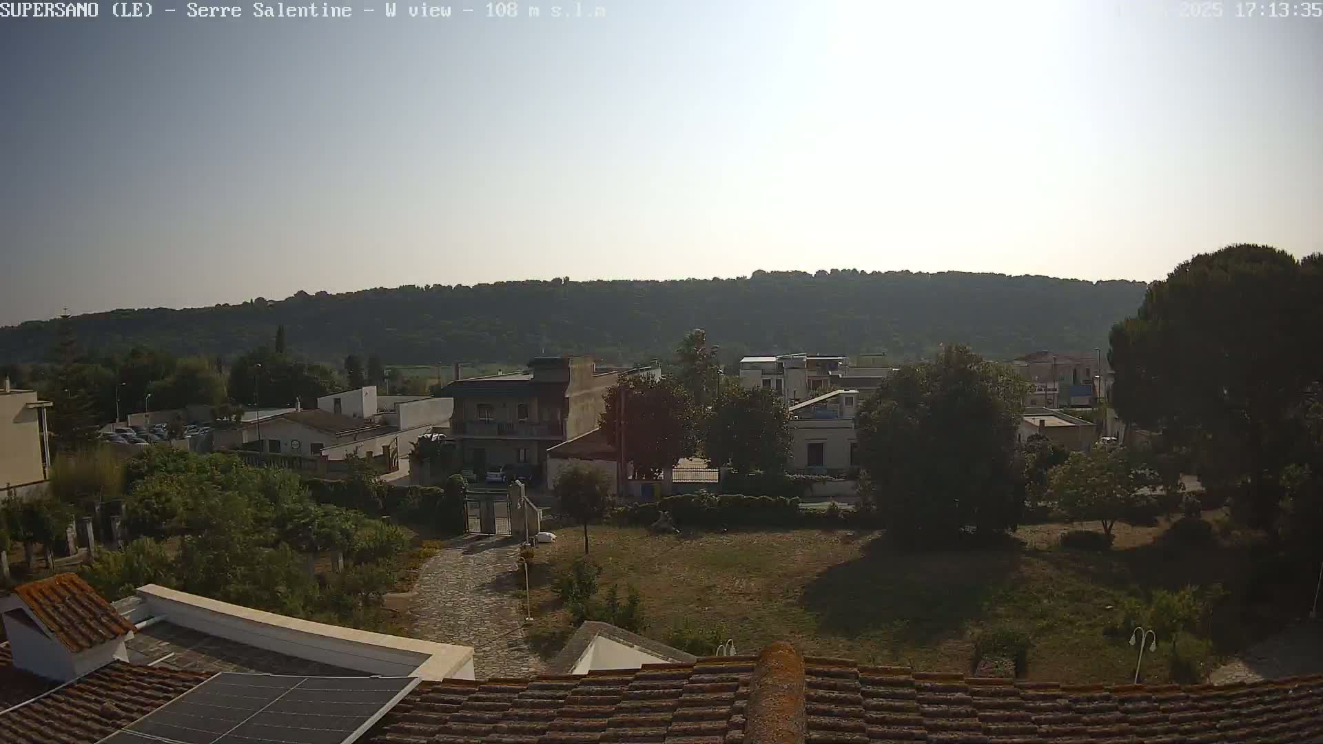 A sunny view of a residential area with low buildings and lush green trees, set against a backdrop of rolling hills.