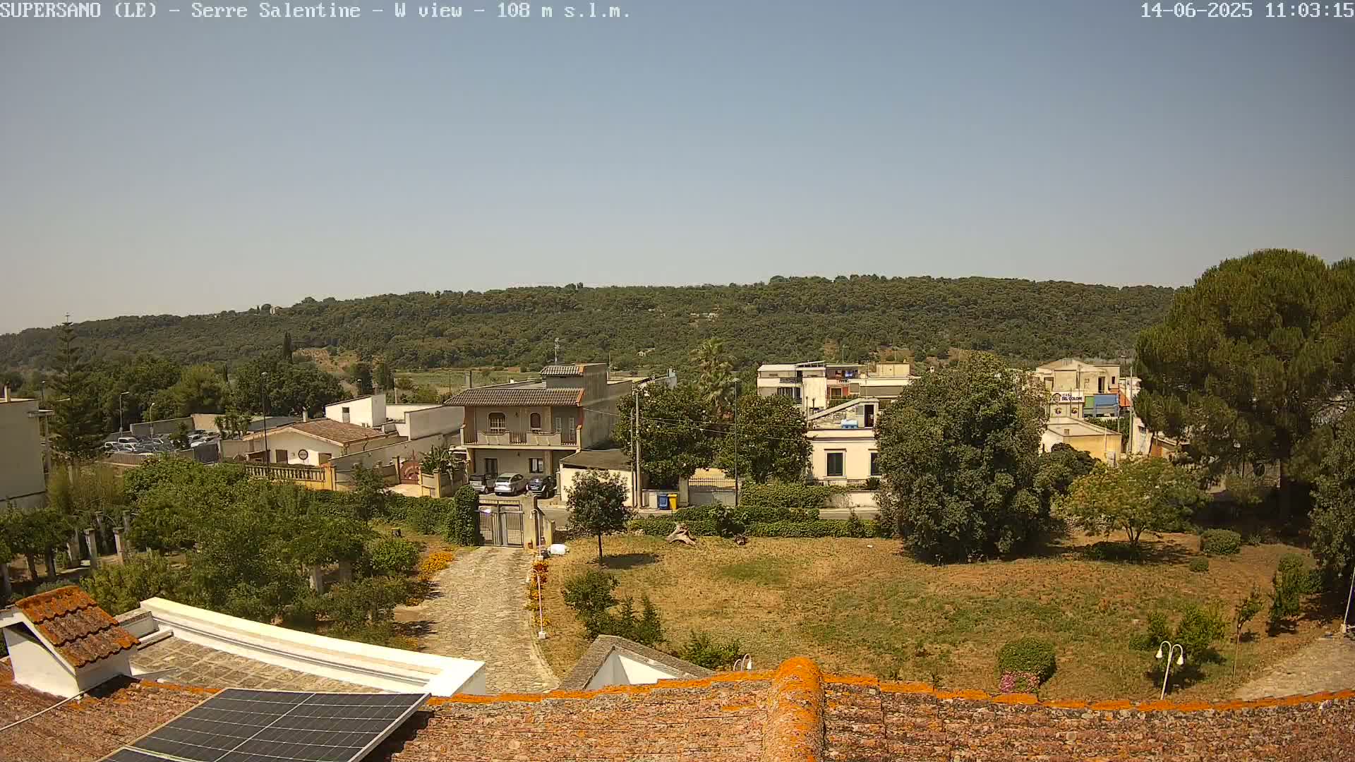 A sunny view of a residential area with houses, trees, and a hill in the background, seen from a rooftop with solar panels.