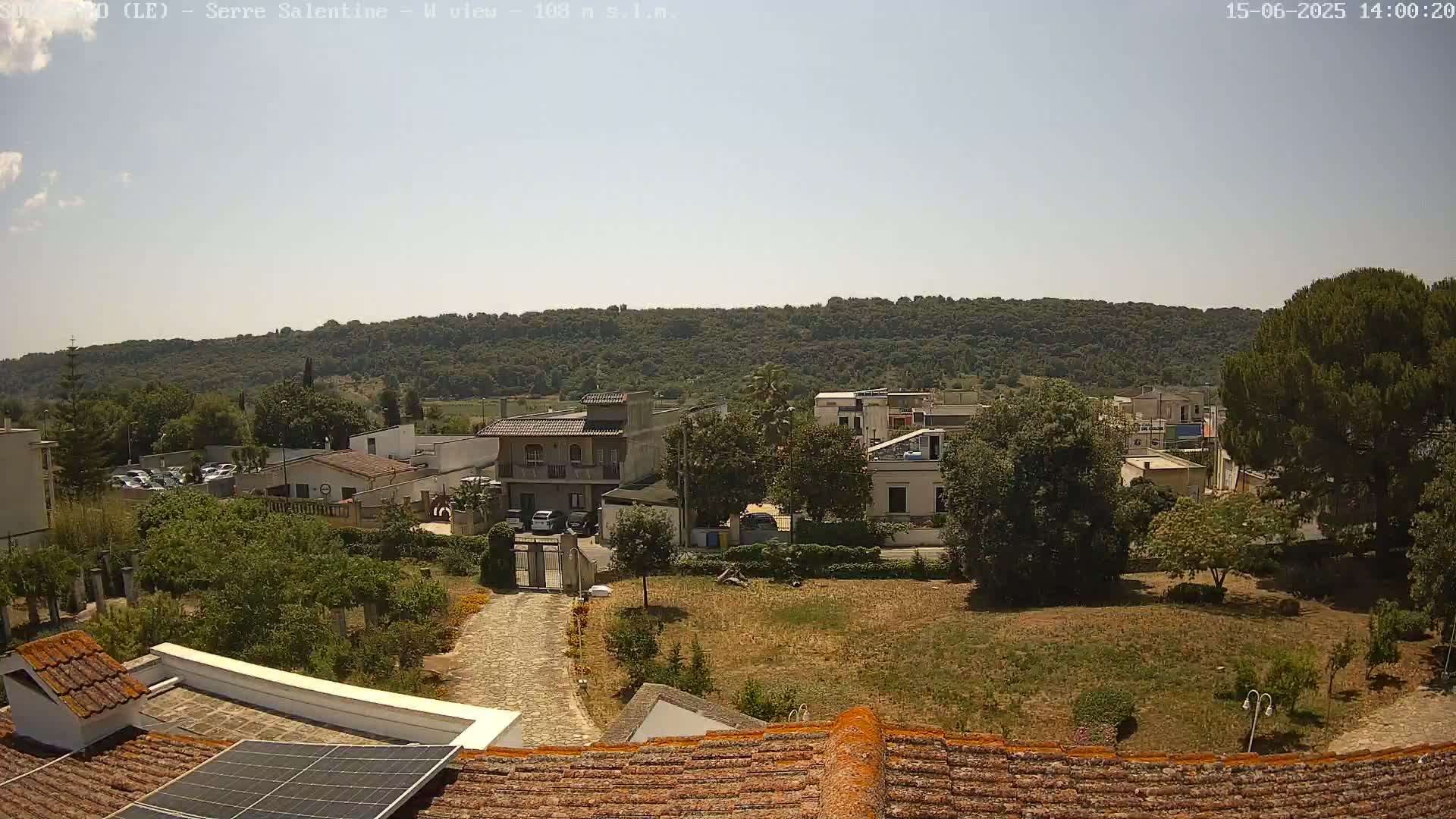 A high-angle, long shot on a sunny day shows a residential area with houses and trees, backed by a hill covered in dense vegetation.
