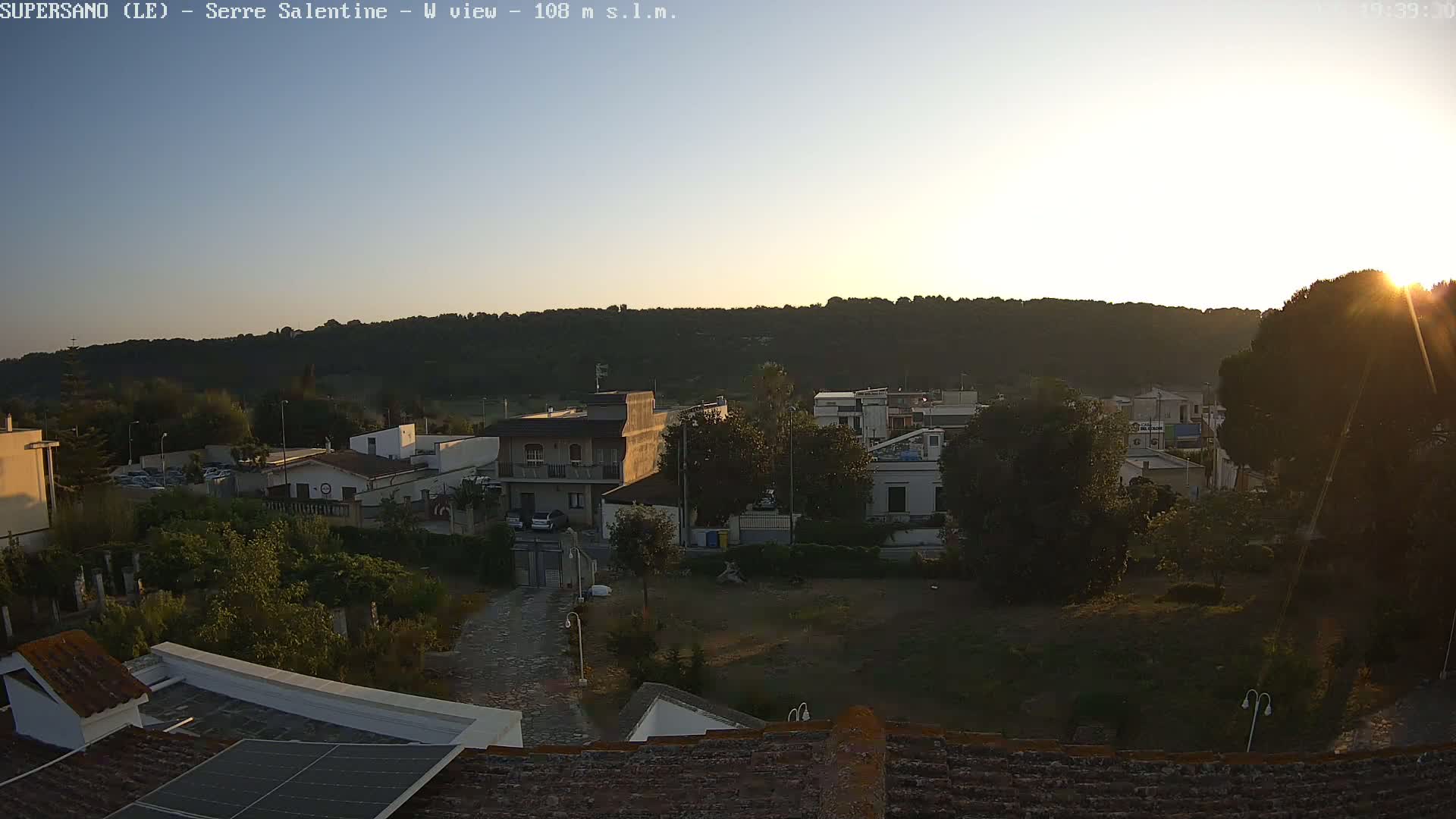 A sunlit view of a residential area nestled against a hill, with the setting sun casting a warm glow over the scene.