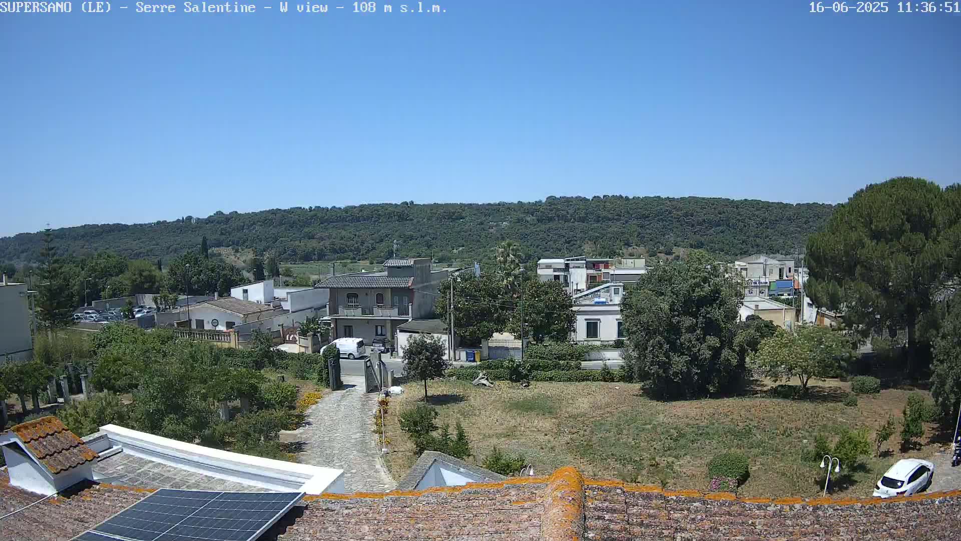 A high-angle, long shot on a clear, sunny day shows a residential area nestled against a verdant hillside, with a stone driveway leading to a house featuring a solar panel on its roof.
