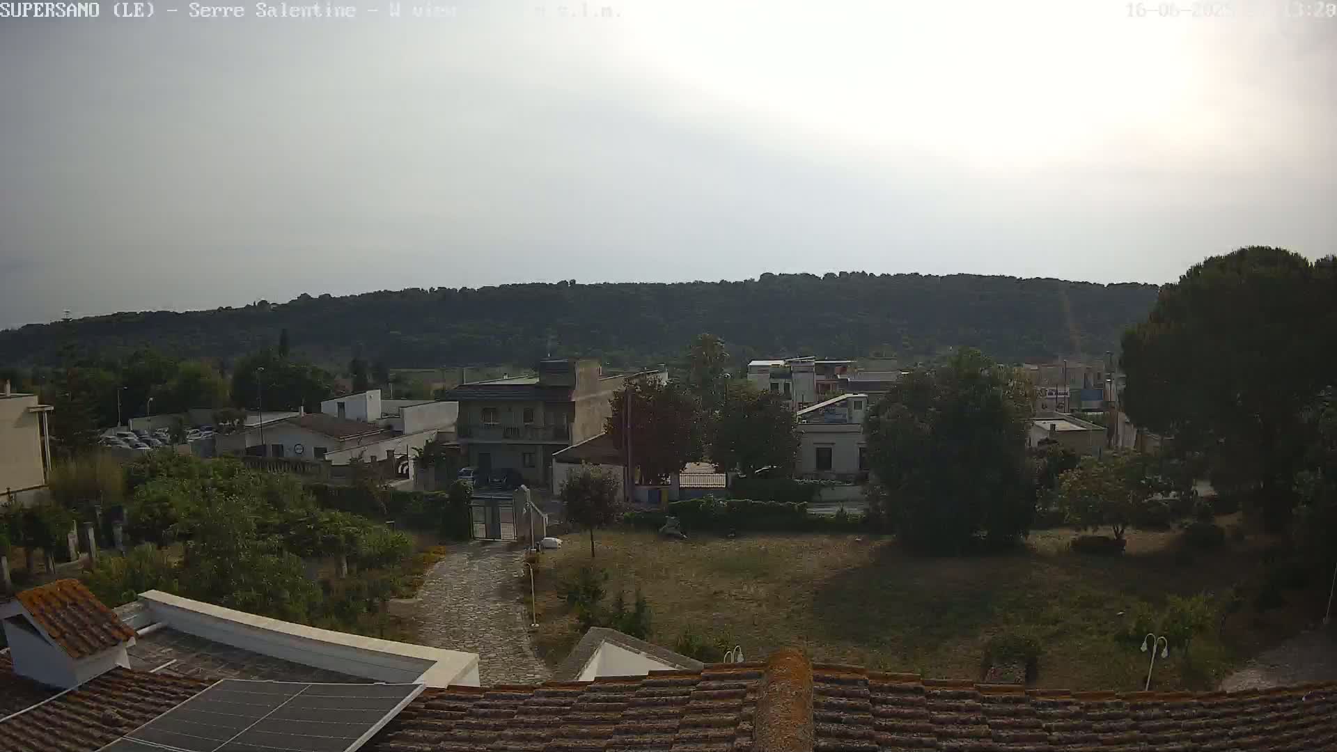 A hazy, sunny day reveals a view of a residential area nestled against a backdrop of rolling green hills, seen from a rooftop with solar panels.