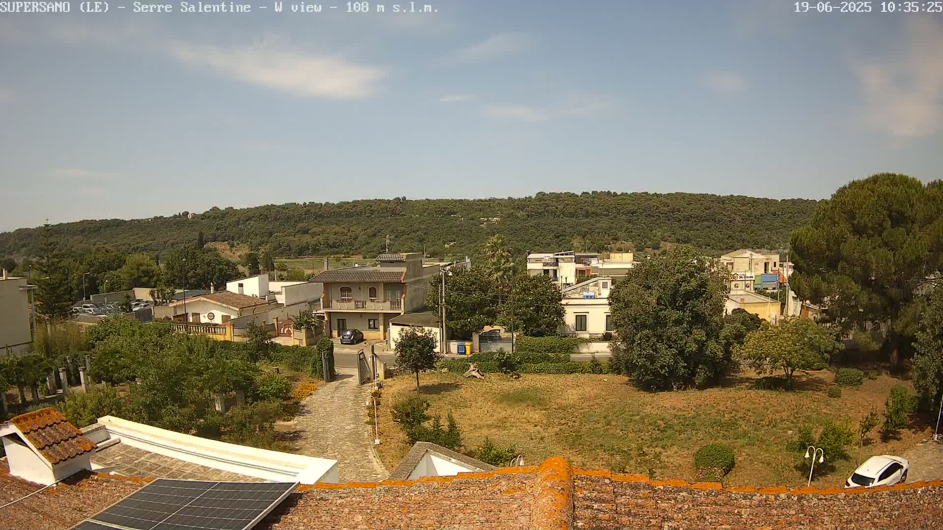 A clear, sunny day shows a view of a residential area with houses, trees, and a paved pathway, all nestled below a tree-covered hillside.