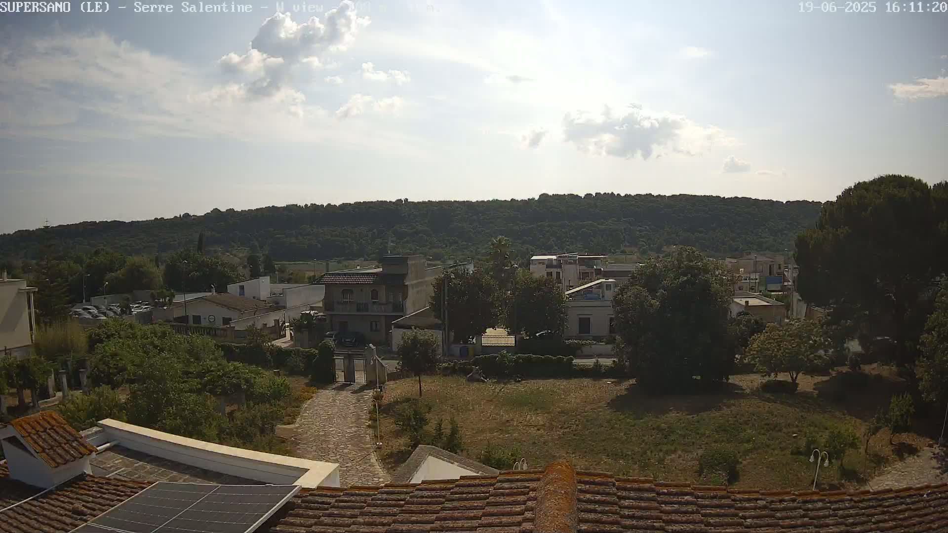 A partly sunny day overlooks a residential area with low buildings, lush green trees, and a hillside in the background.