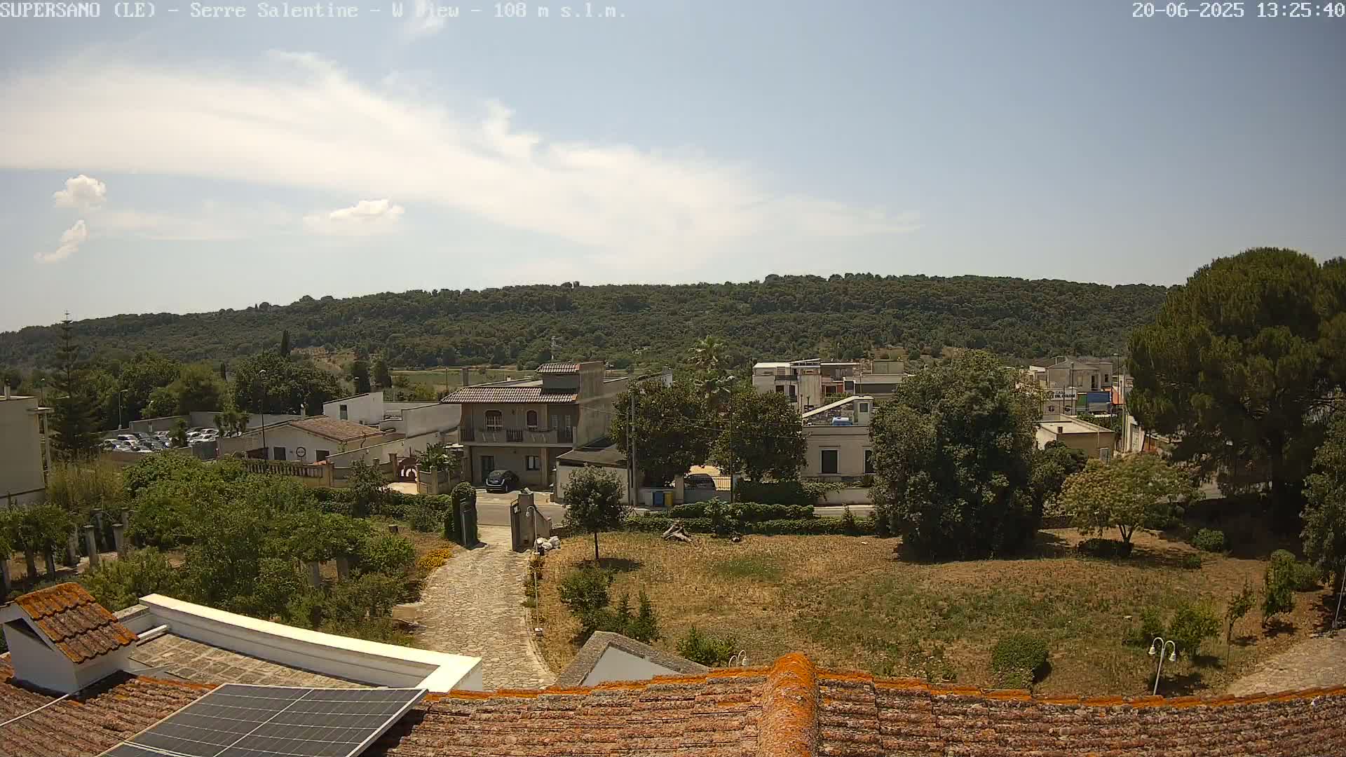 A mostly sunny day reveals a small town nestled at the base of a tree-covered hill, viewed from a rooftop with a solar panel.