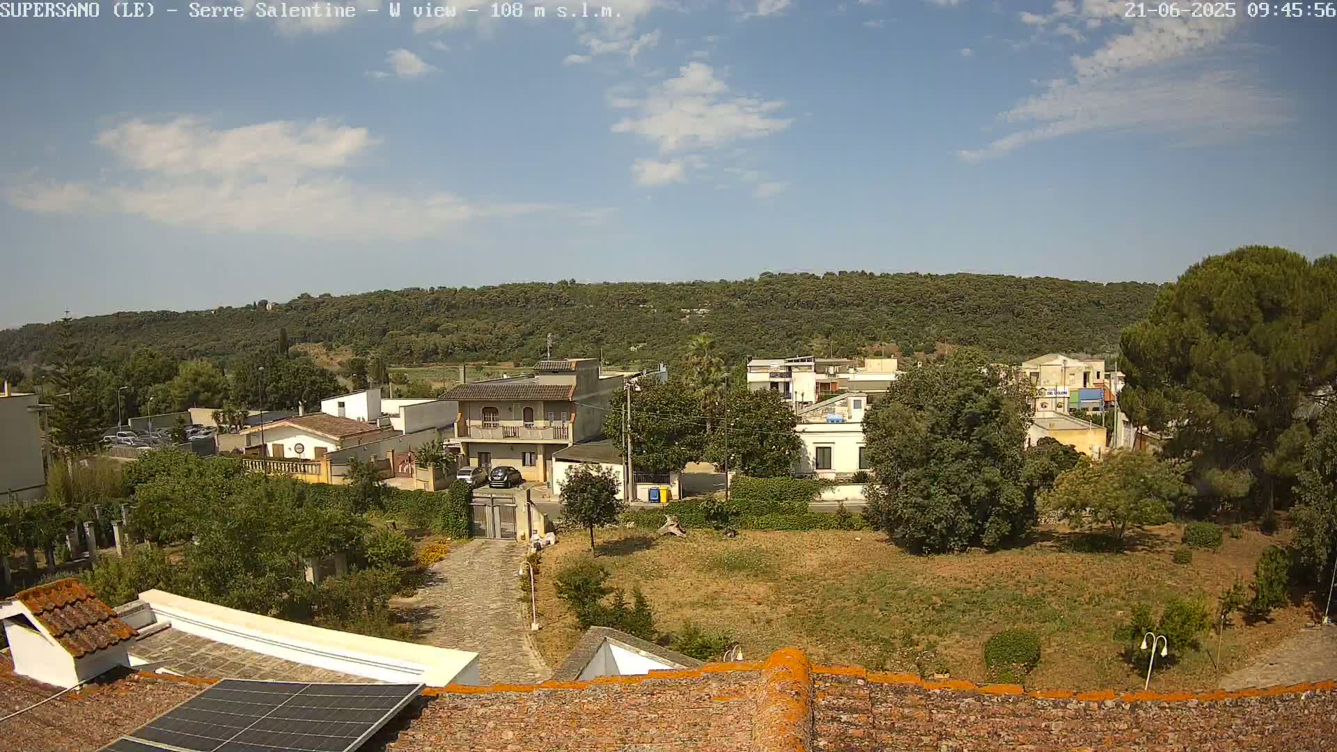 A sunny day reveals a residential area nestled below a verdant hillside, with a paved pathway leading to a building equipped with solar panels.